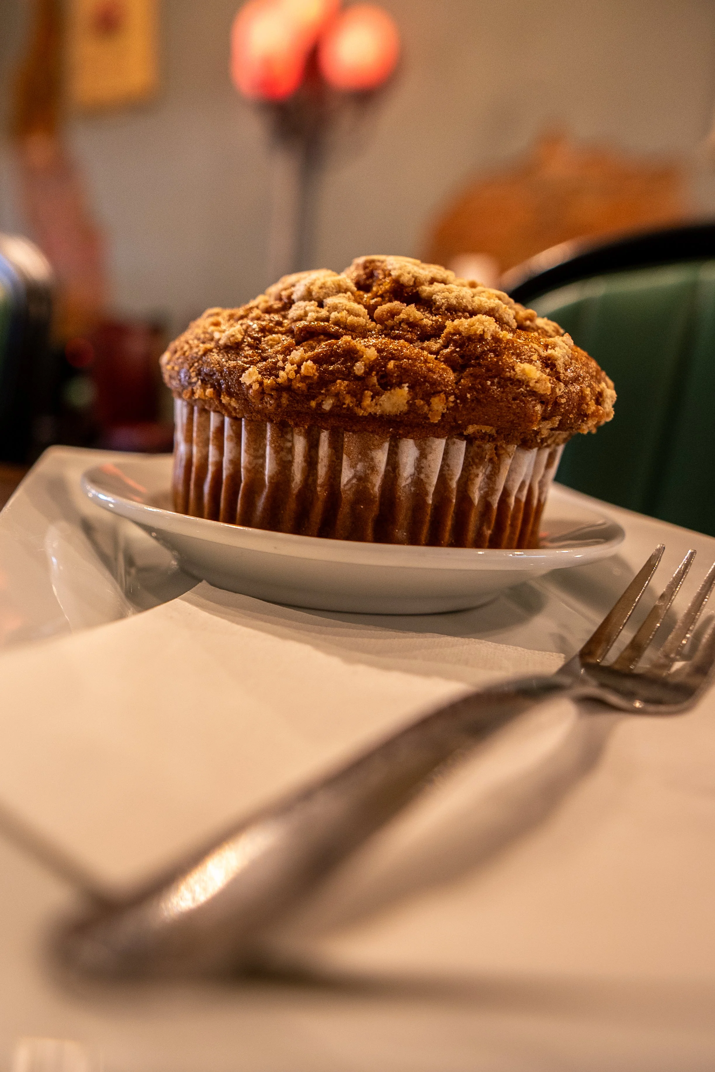 Close-up of a muffin with a crumbly topping on a white plate, with a fork in the foreground and a blurred background including a lamp and decorative items.