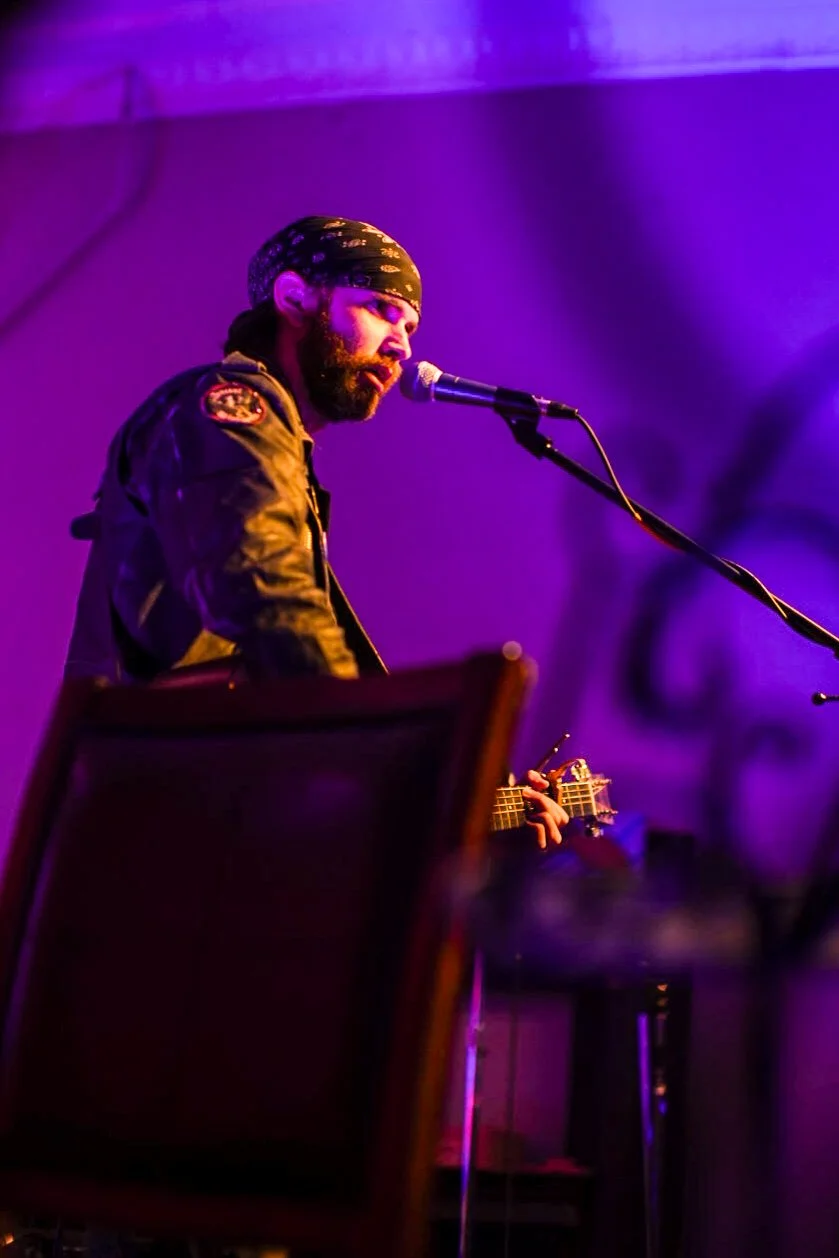 A bearded musician with a bandana on his head playing guitar and singing into a microphone on stage bathed in purple and blue lighting.