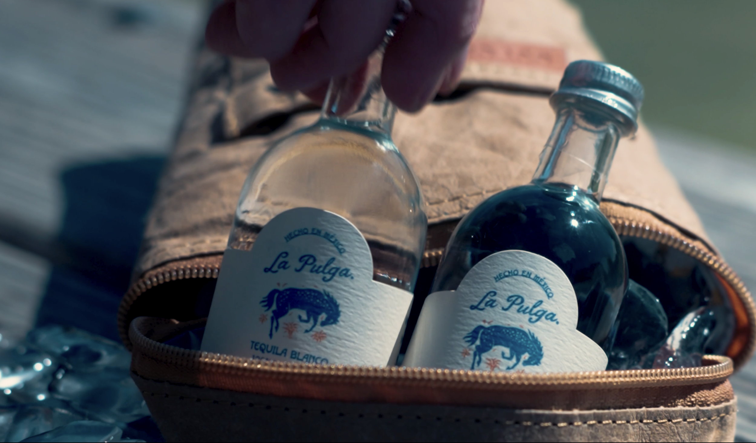 Close-up of a hand reaching into a tan bag containing several small bottles of tequila with blue and white labels labeled 'La Pulga' and an image of a blue flea, with a zippered edge of the bag visible.