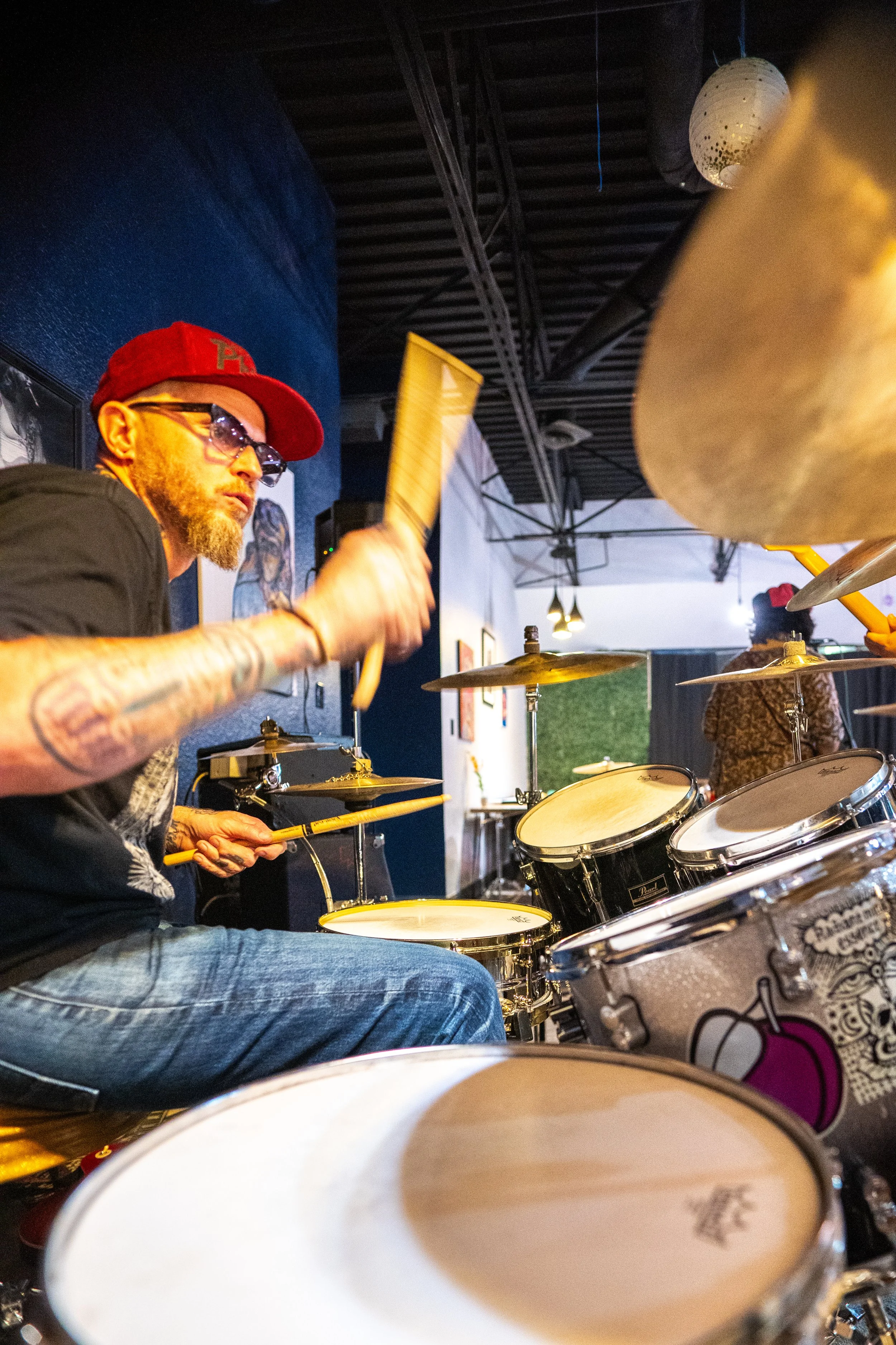 A man wearing sunglasses and a red cap playing a drum set in a music venue, with other performers and artwork in the background.