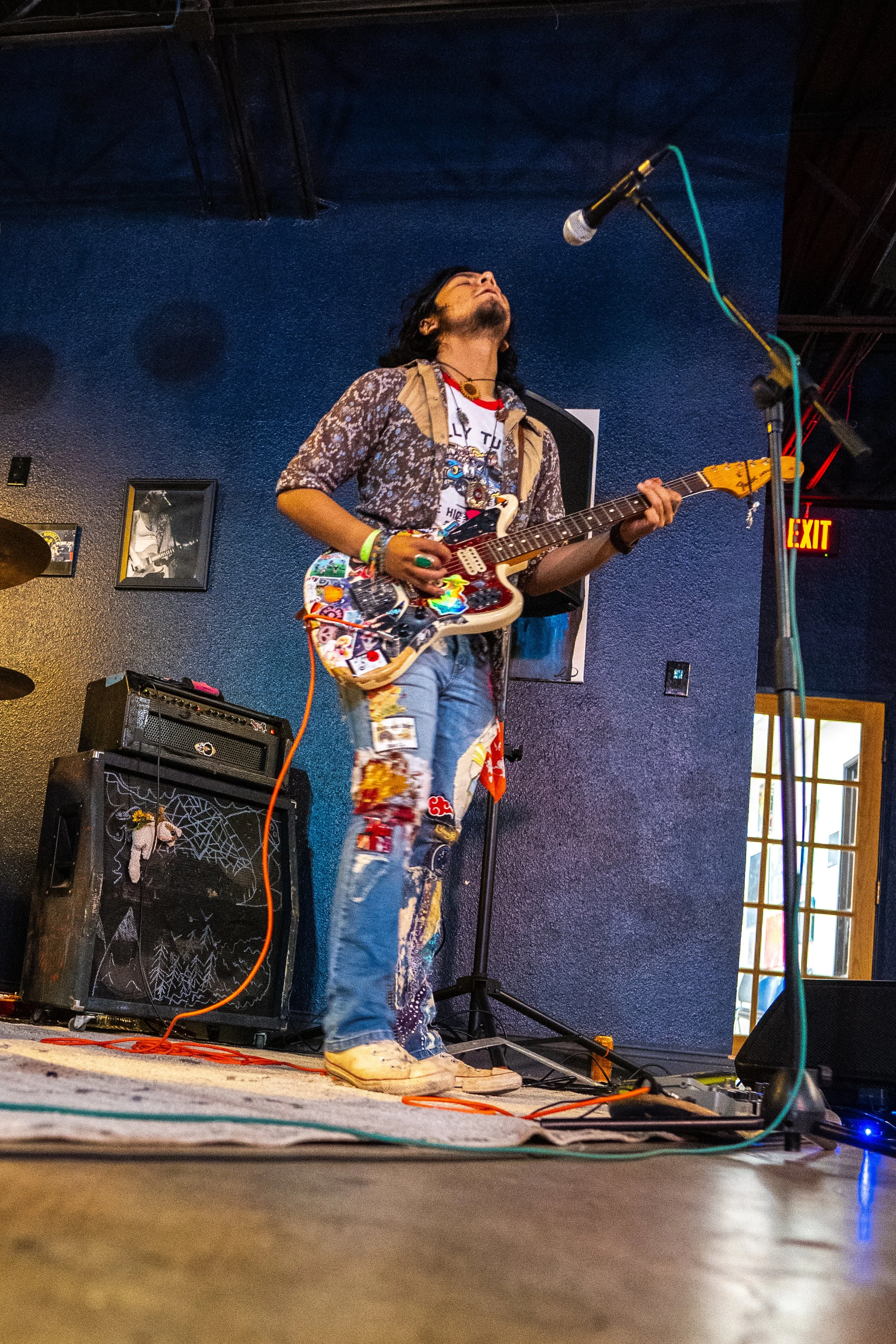 A musician with long dark hair, wearing glasses, a patterned shirt, distressed jeans, and sneakers, is playing an electric guitar on stage with a microphone in front. The background features a dark blue wall, framed pictures, and stage equipment, wit