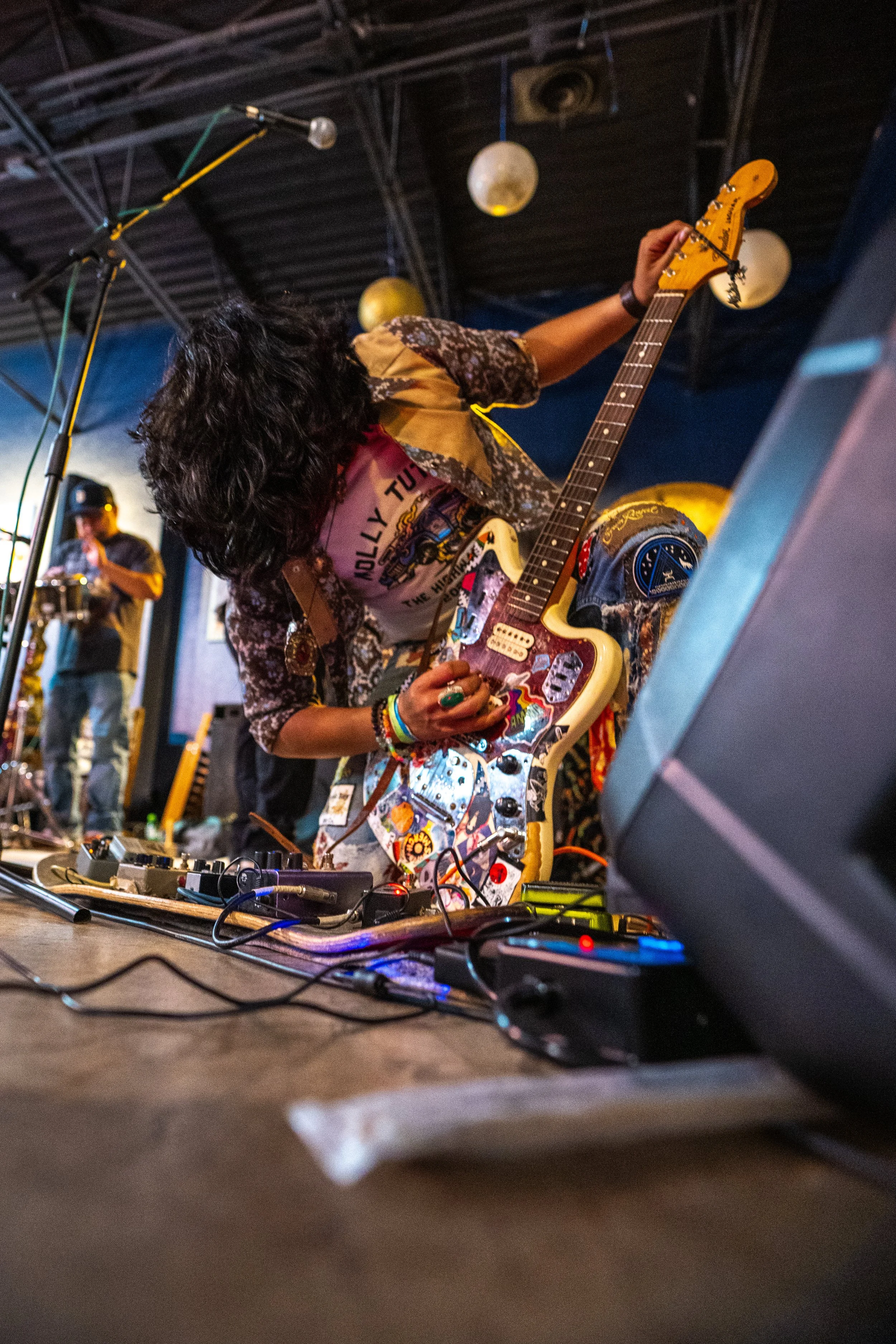 Musician playing an electric guitar on stage with effects pedals and a laptop in front, under dim lighting with hanging paper lanterns overhead.