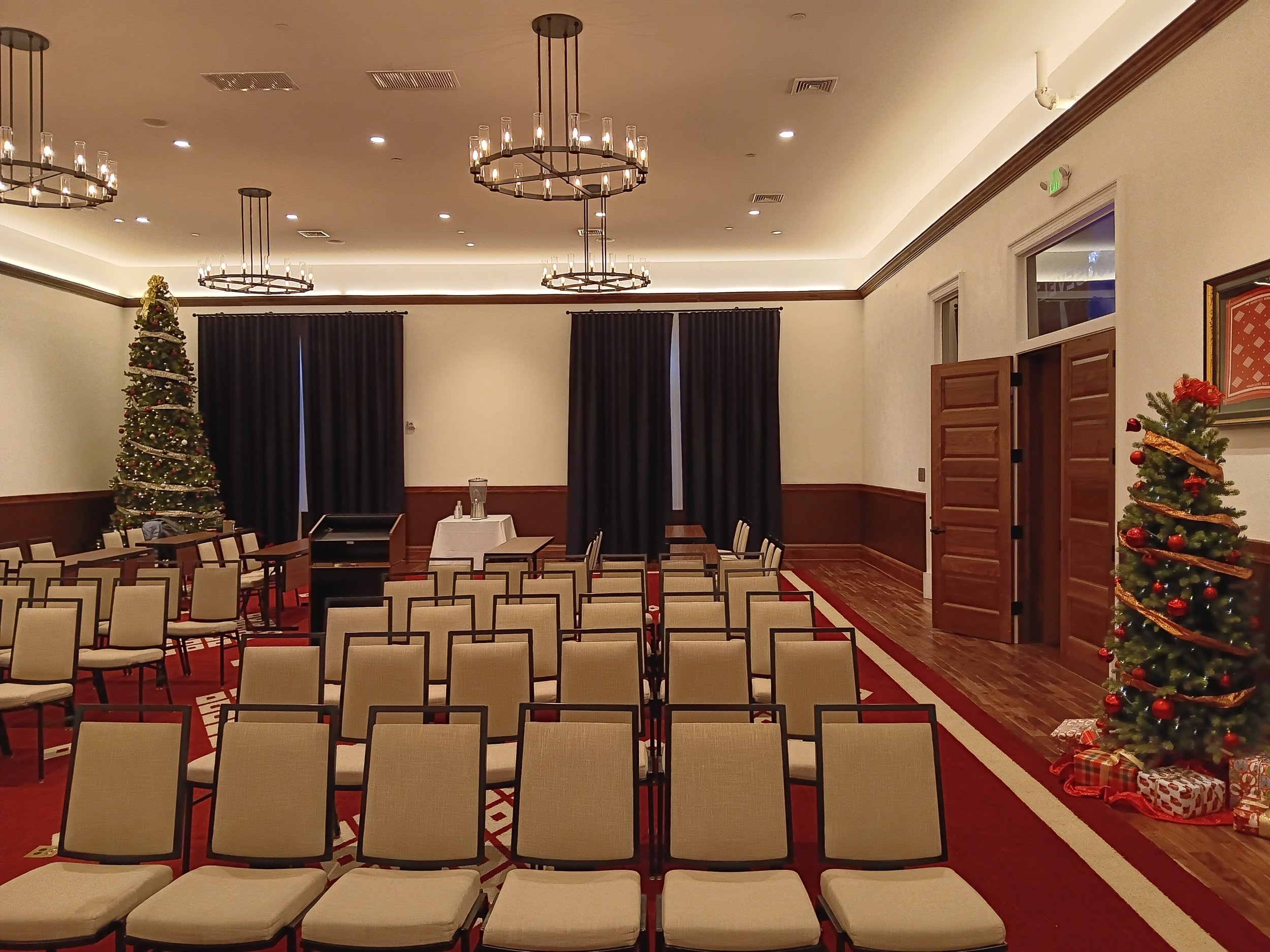Empty conference room decorated for Christmas with two Christmas trees, chairs arranged in rows, a table with water pitchers, and black curtains.