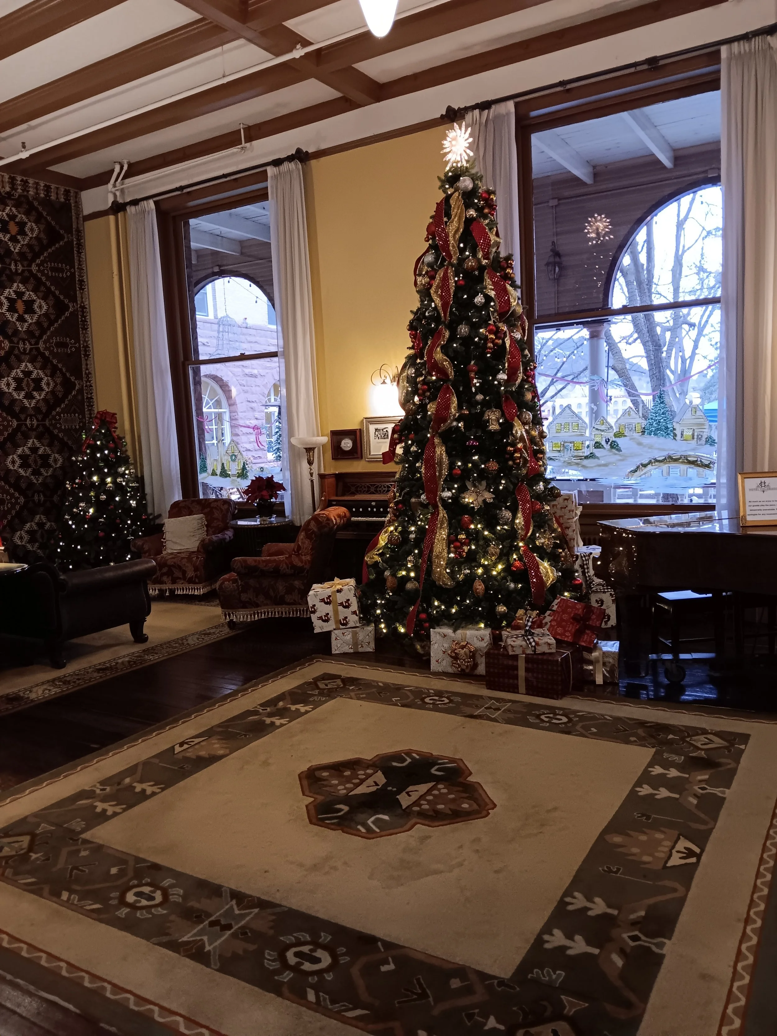 Christmas tree decorated with ornaments, ribbons, and lights, surrounded by wrapped presents, in a cozy room with large windows and holiday decorations.