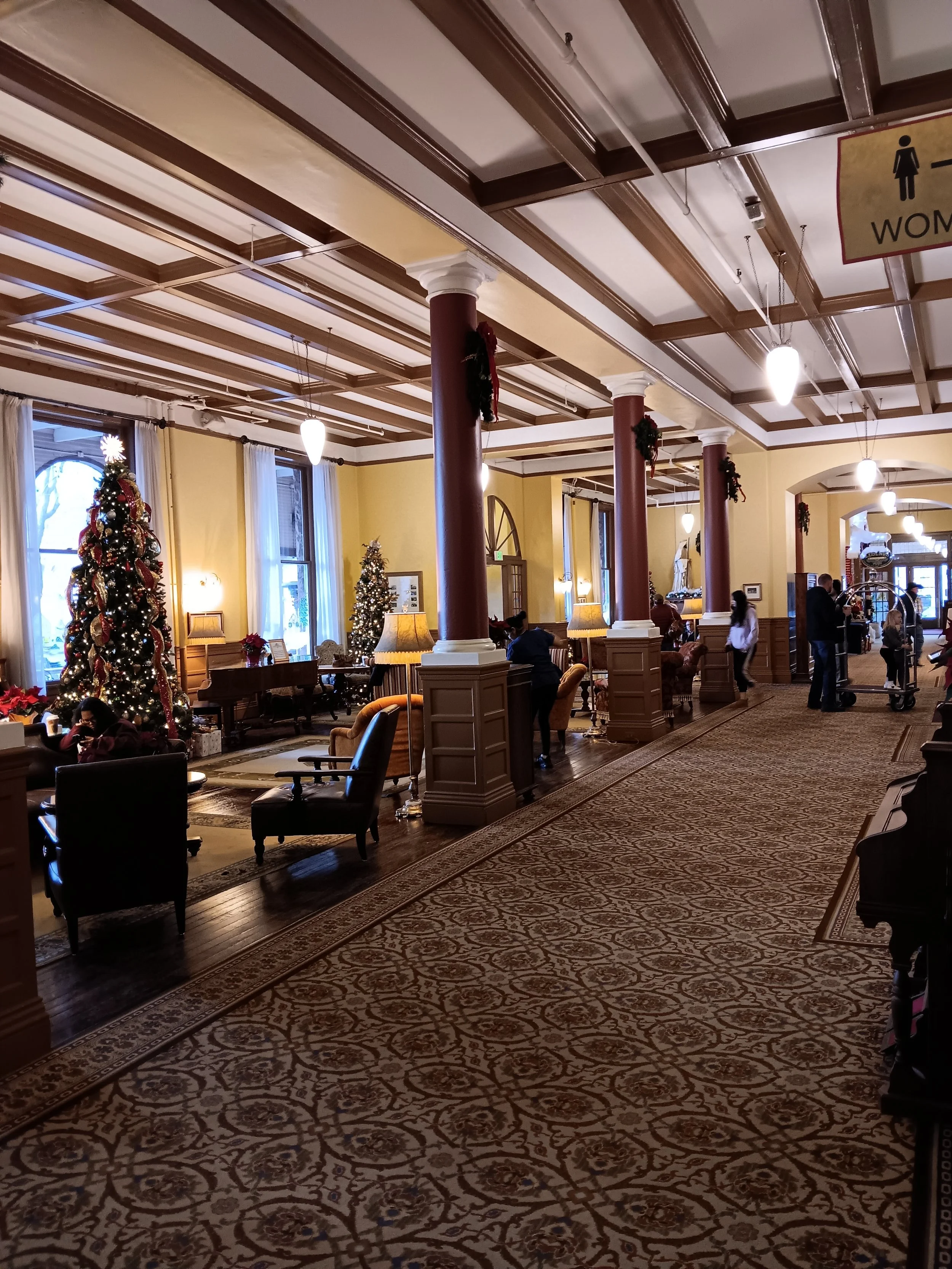 Lobby decorated for Christmas with tall decorated Christmas trees, wreaths, and holiday lights. People sitting and standing, some by tables and lamps, in a spacious, elegant room with large windows, yellow walls, and ornate ceiling beams.
