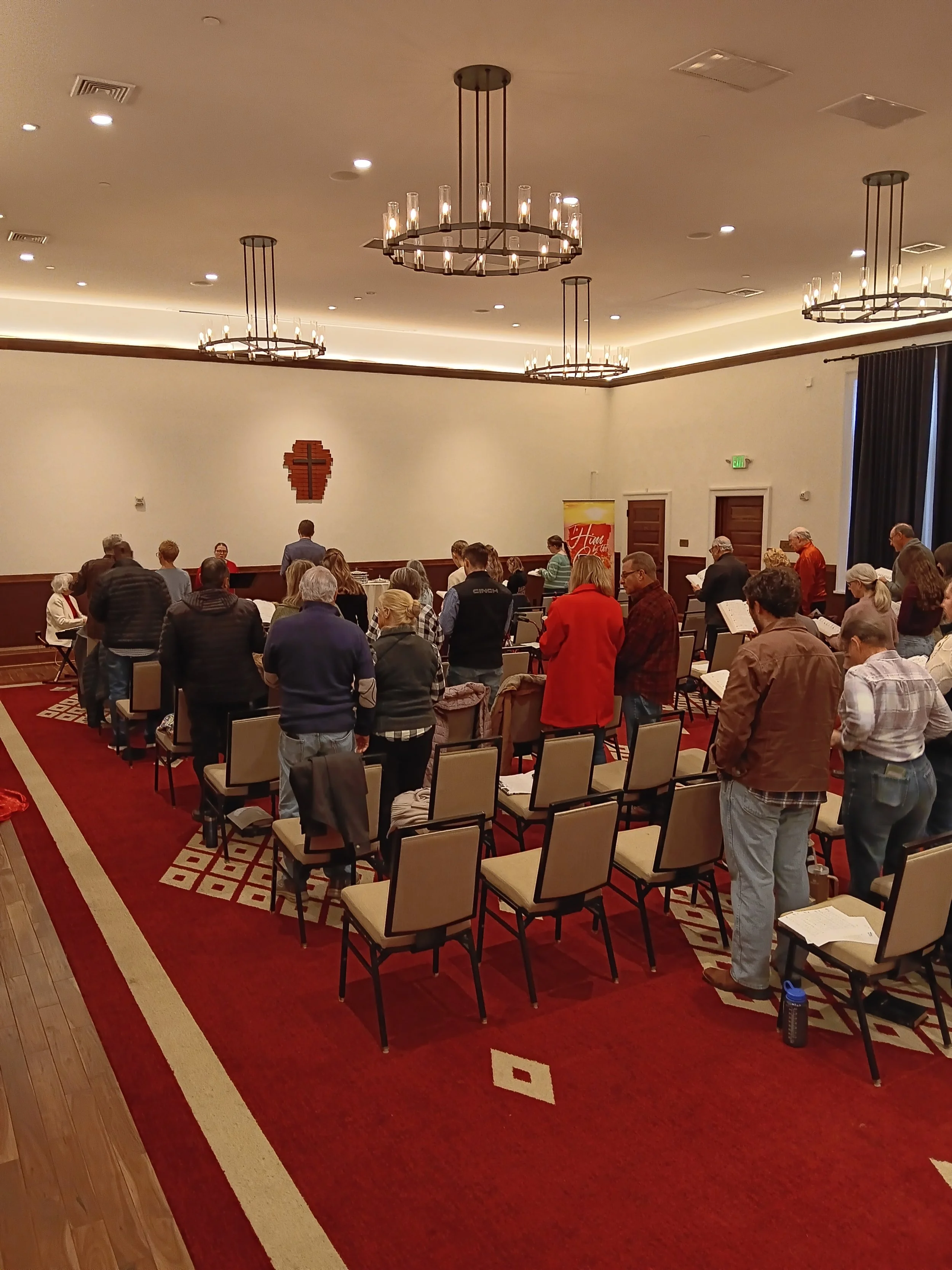 A group of people participating in a prayer or worship service in a church, with some standing and some sitting, facing the altar area at the front of the room.