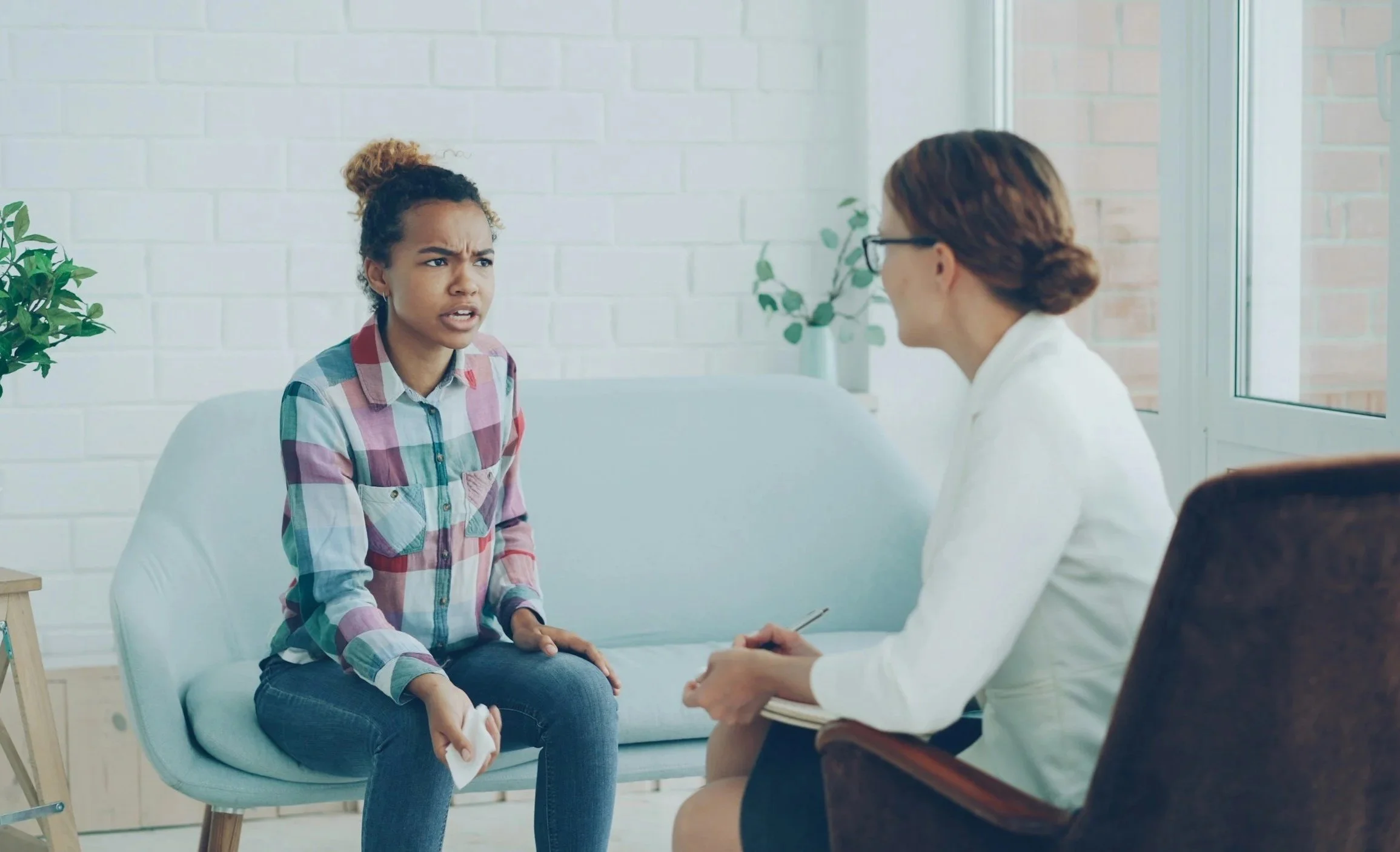 A young woman with curly hair in a bun wearing a colorful plaid shirt sits on a light blue couch, appearing upset. A professional woman with brown hair in a bun, wearing glasses and a white blazer, sits in an armchair facing her, taking notes and listening attentively. They are in a bright room with white brick walls, a window, and plants.