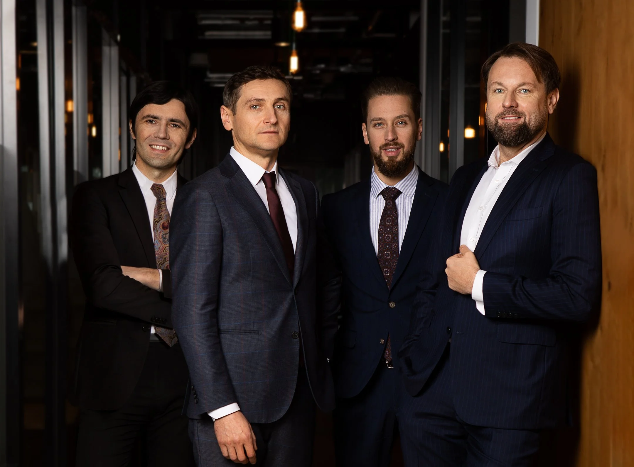 Four men in suits standing in a hallway, looking at the camera.