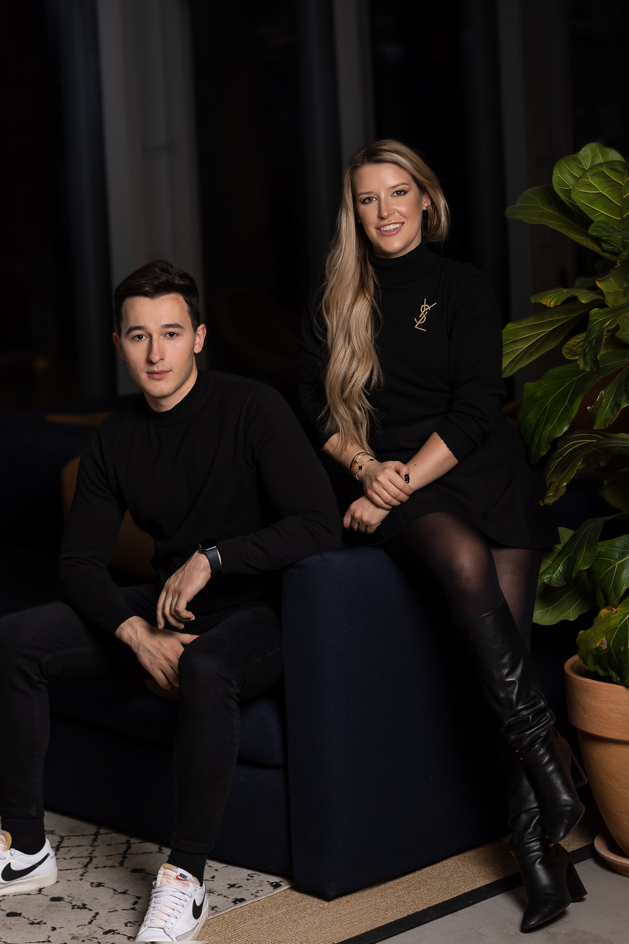 A man and a woman pose on a dark couch in a modern indoor setting with large green indoor plants.