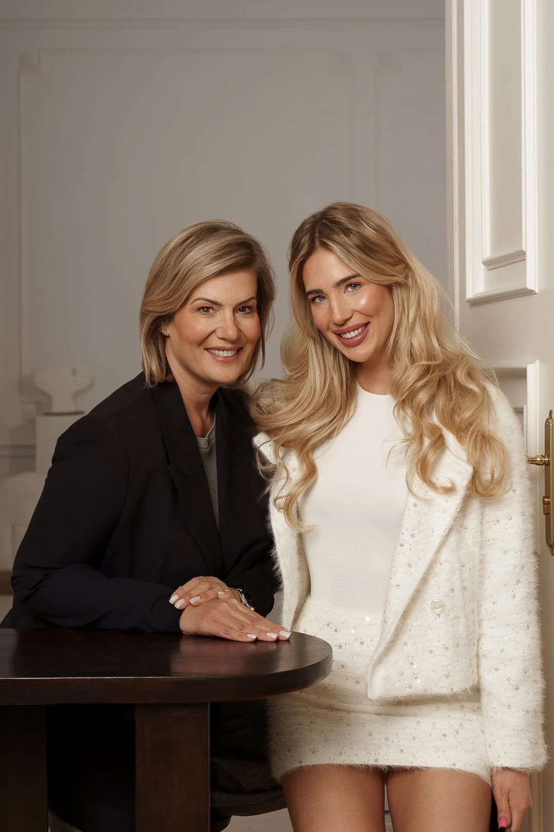 A woman with shoulder-length blonde hair and a woman with long, wavy blonde hair smiling together indoors.