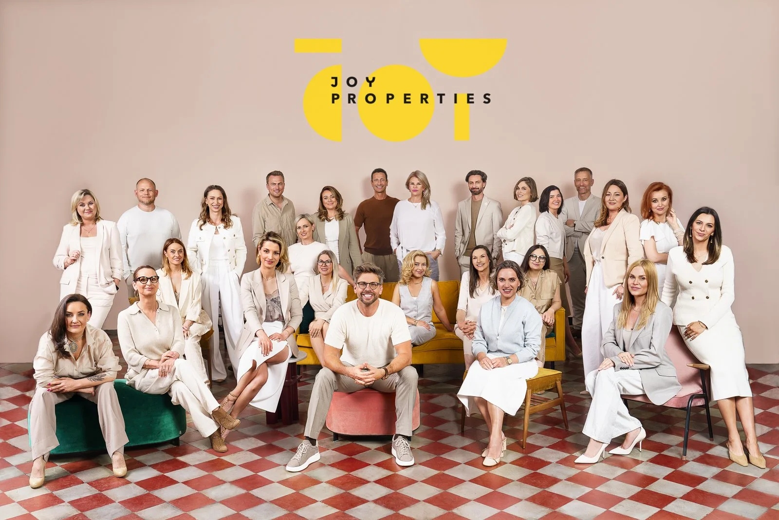 A large group of men and women dressed in neutral tones, posing in front of a beige background with the words 'Joy of Properties' in stylized yellow and black font at the top. Some are seated on colorful chairs and couches, while others are standing 