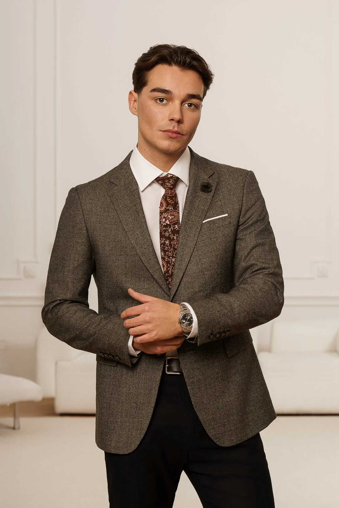 A young man in a brown blazer, white shirt, patterned tie, and black pants standing in a room with white walls and light-colored furniture.