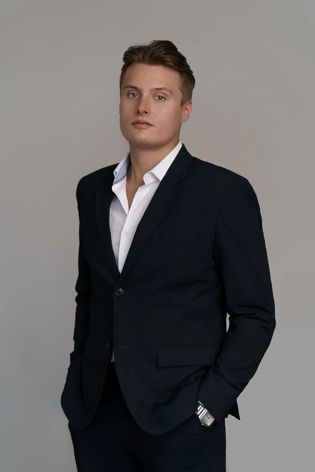 A young man with light skin, wearing a white dress shirt and a black blazer, standing against a plain gray background with his left hand in his pocket and looking at the camera.