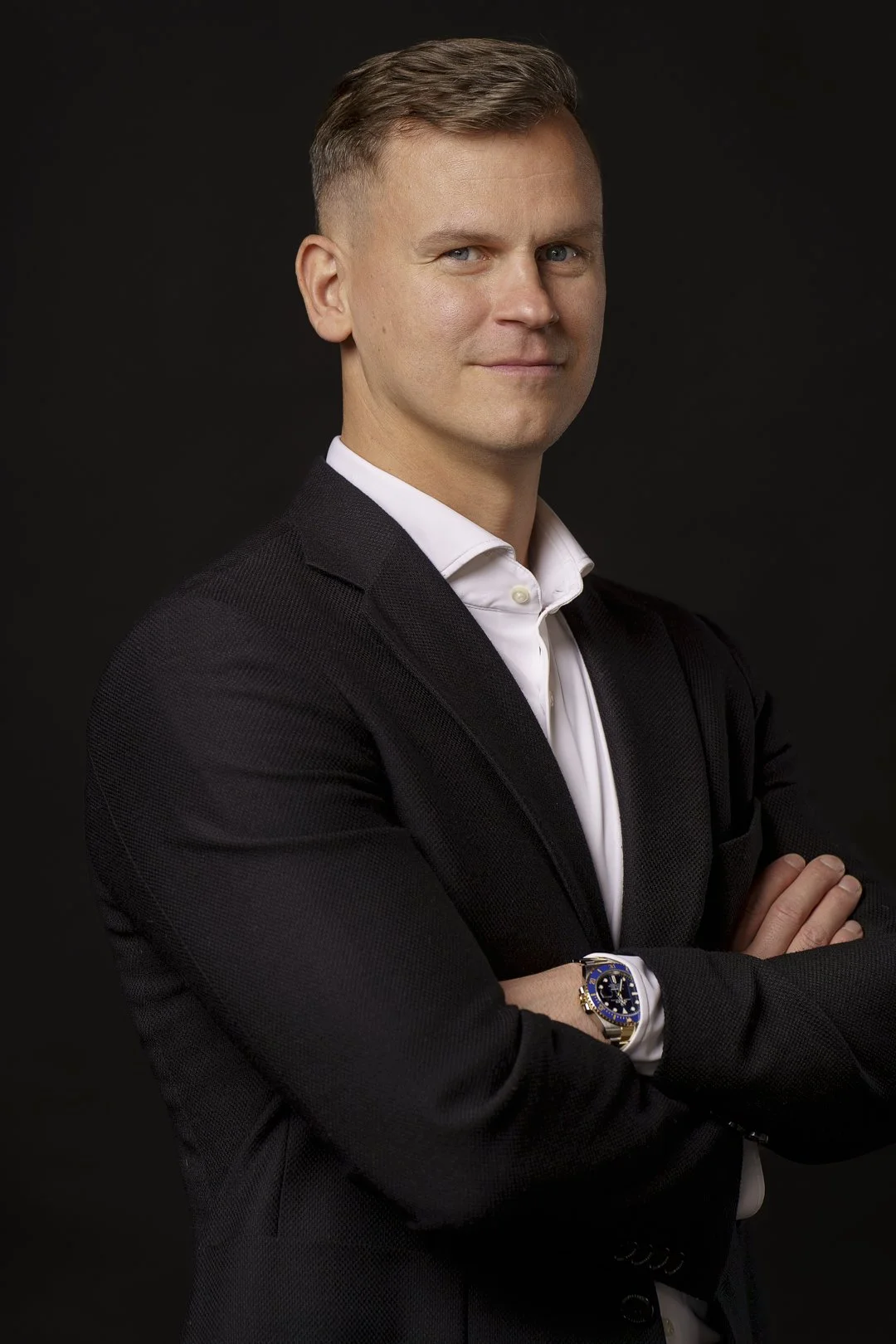 A man with short light brown hair, wearing a dark blazer and white shirt, standing with arms crossed against a black background.