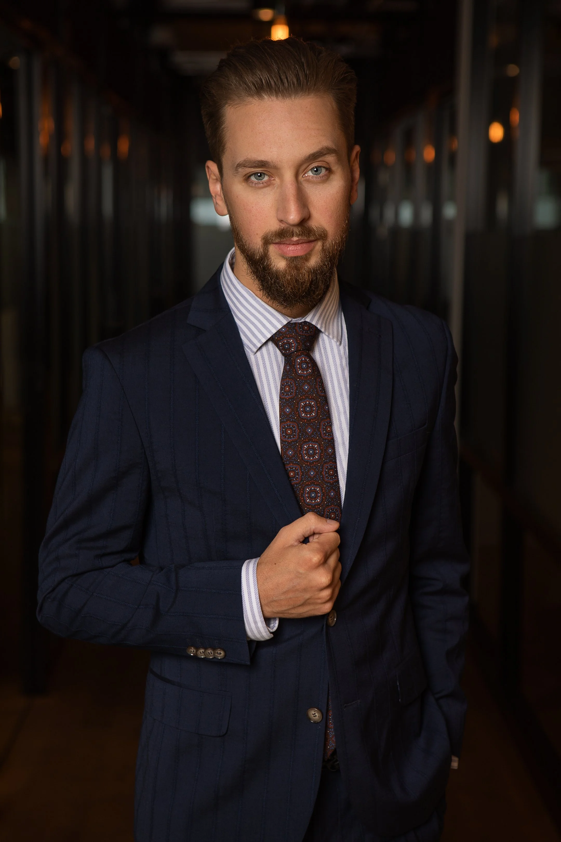 A confident man with a beard, blue eyes, and styled hair in a dark navy suit, striped shirt, and patterned tie, standing in an indoor setting with warm lighting and modern design elements.
