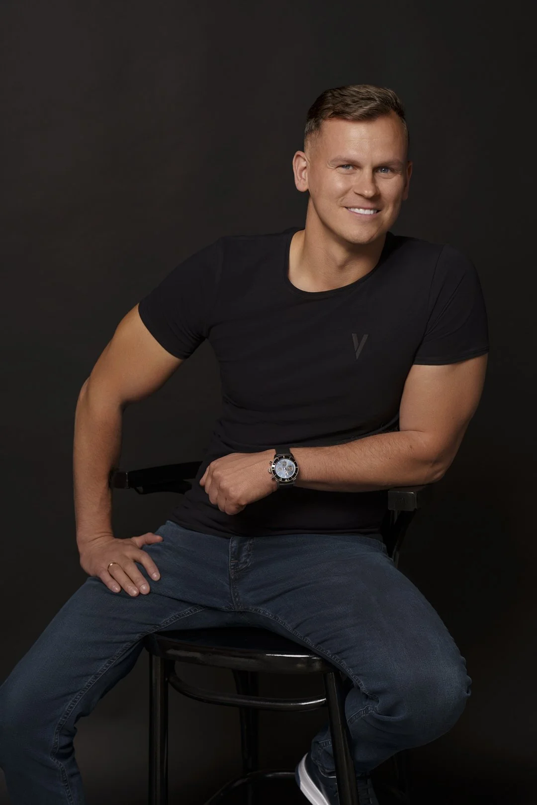 A young man with short hair, wearing a black t-shirt and blue jeans, is sitting on a black stool against a dark background. He is smiling and looking at the camera, with his right hand resting on his knee and his left hand on his wrist.