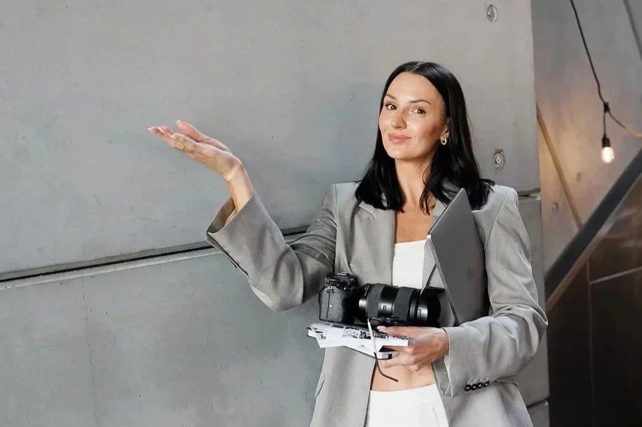 A woman with black hair, wearing a gray blazer, holding a camera, a laptop, and some papers, standing against a gray wall with a string of lights.
