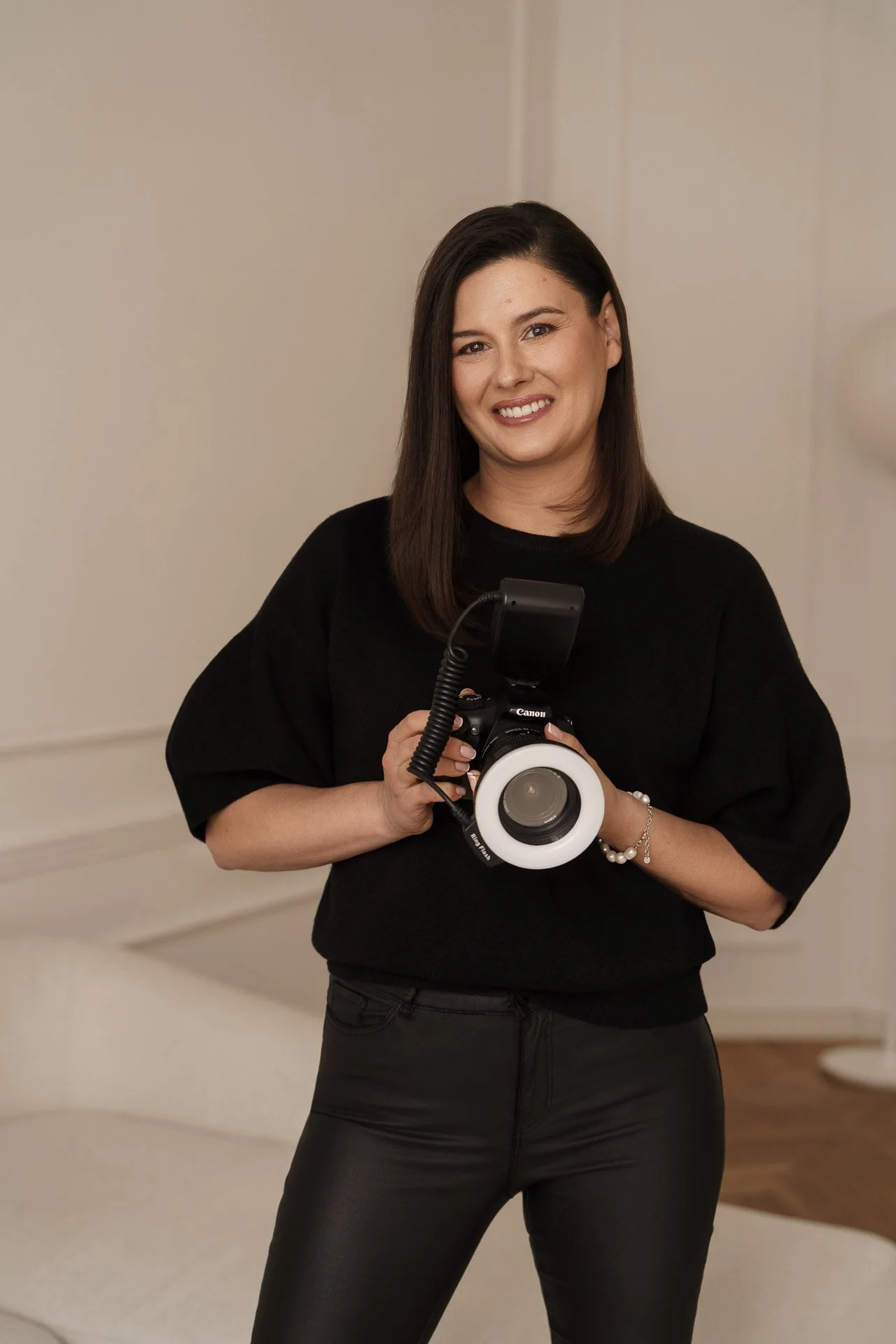 A woman with shoulder-length dark hair, wearing a black top and black pants, smiling and holding a professional camera with a large white lens in an indoor setting.