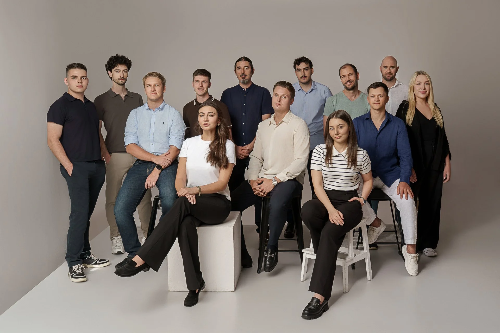 Group of fifteen young adults posing for a photo in a studio with plain background, some sitting on chairs or stools, others standing behind.