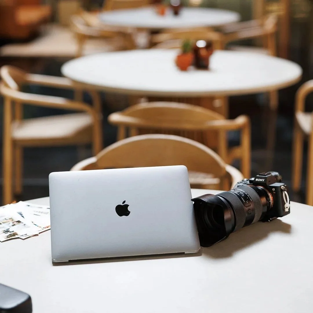A closed silver MacBook, a professional video camera with a large lens, and some photographs on a white table in a bright, outdoor setting with wooden chairs and tables in the background.