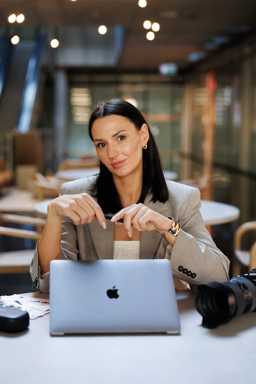 A woman with dark hair, wearing a beige blazer, sitting at a table with a closed MacBook, a camera, and papers, in a modern, well-lit cafe or workspace.