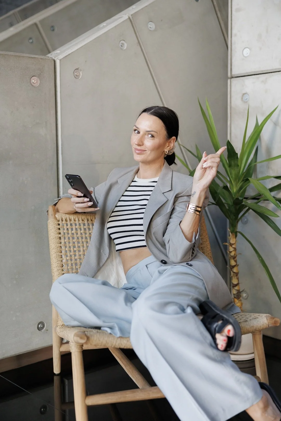 A woman in a gray blazer and striped crop top sitting in a wicker chair, holding a smartphone, smiling with a finger pointing upward, next to a green potted plant in an indoor modern setting.