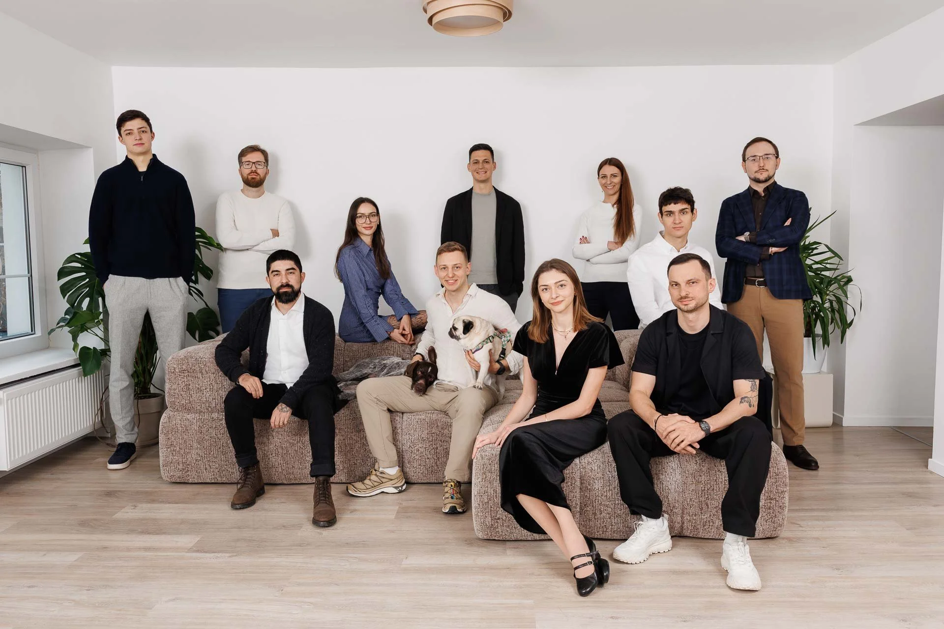 Group of twelve young adults posing in a bright, modern living room with white walls and wooden flooring.
