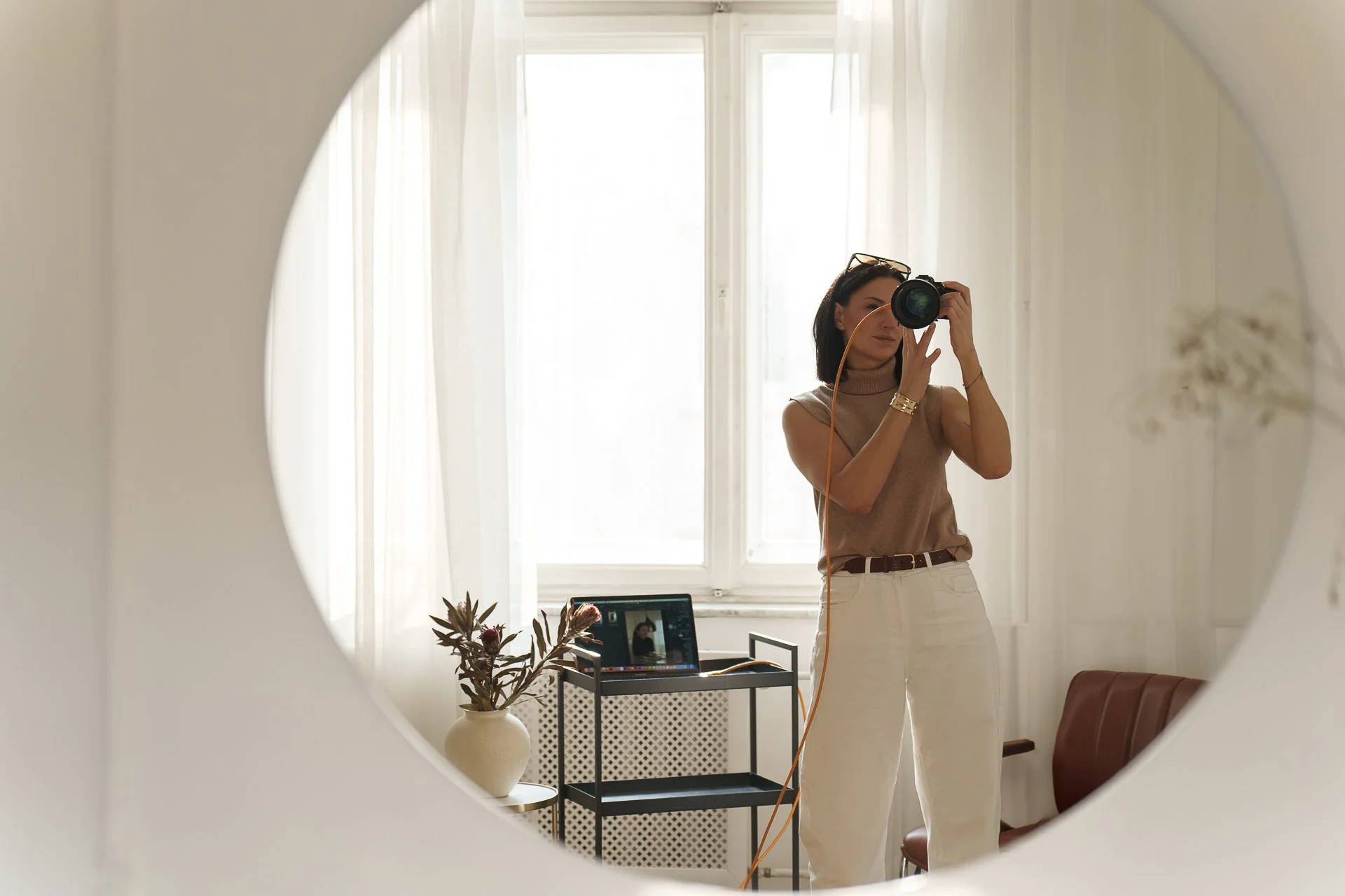 Woman taking a photo of herself with a camera in a mirror, in a bright room with large windows, sheer curtains, and a small table with a plant and a laptop