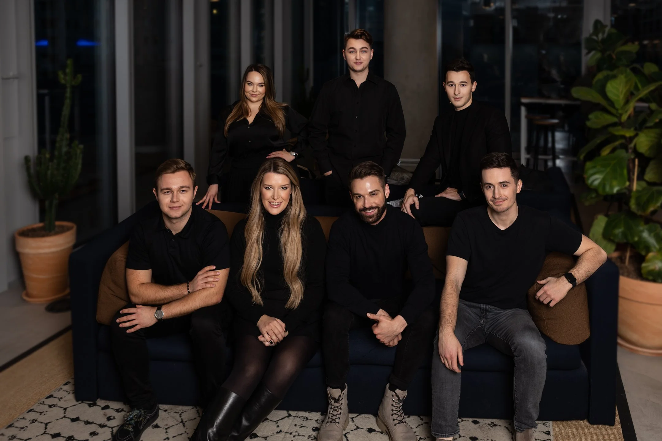 Group of eight young adults gathered in a modern indoor setting, sitting and standing around a sofa, dressed in black, smiling at the camera.