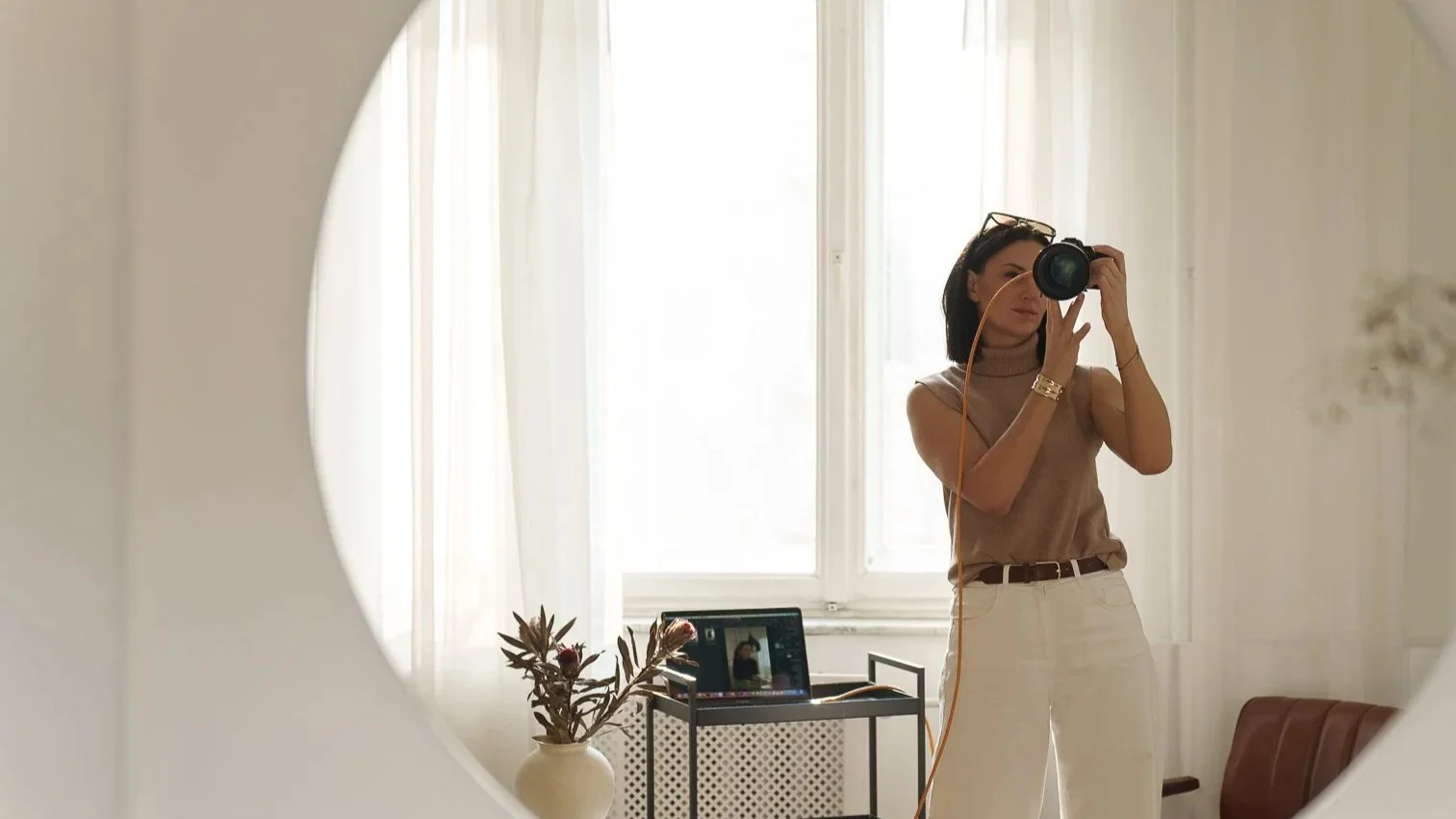 A woman taking a self-portrait with a camera in a mirror, standing in a bright room with curtains, a smartphone on a small table, and a vase with dried plants.