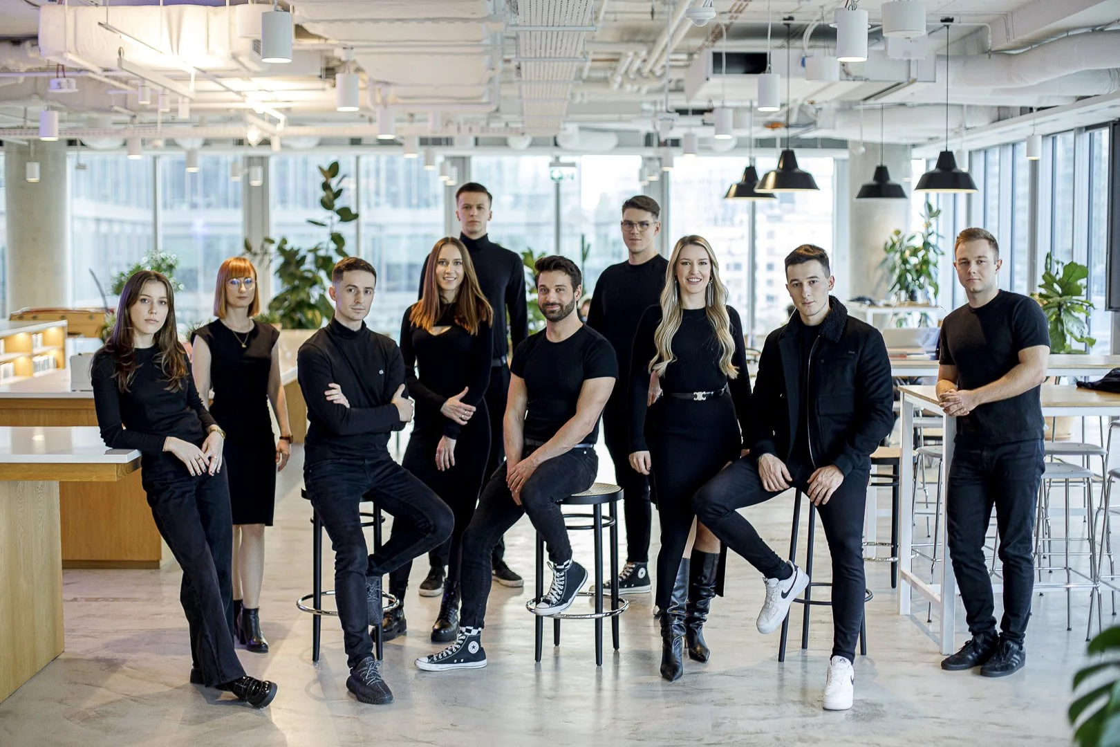 A group of ten diverse young professionals in black attire gathered in a modern office space with large windows, plants, and bright lighting.