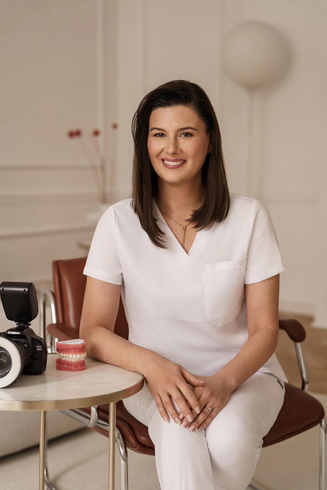A woman in a white medical uniform sitting at a table with a model of teeth and a camera.