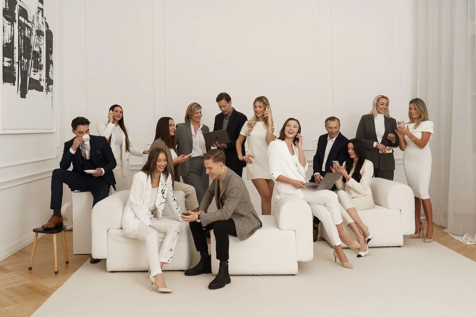 A group of twelve businesspeople in formal attire socializing and working together in a bright, modern office room with white walls and a white couch.