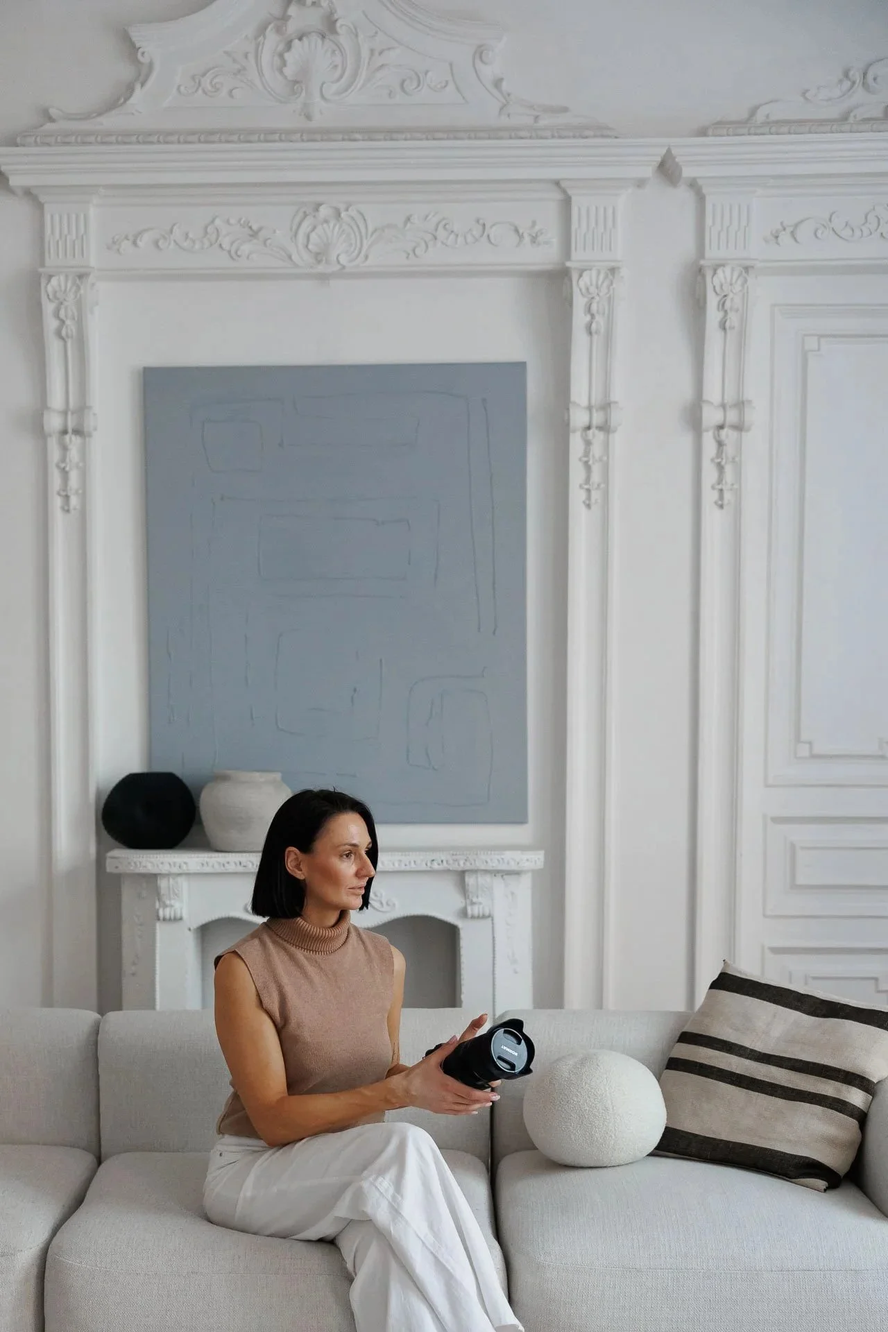 A woman with black hair, wearing a sleeveless tan top and white pants, holding a black camera, seated on a light-colored sofa in a room with ornate white walls and a fireplace, with decorative black and white vases and a striped pillow on the sofa.