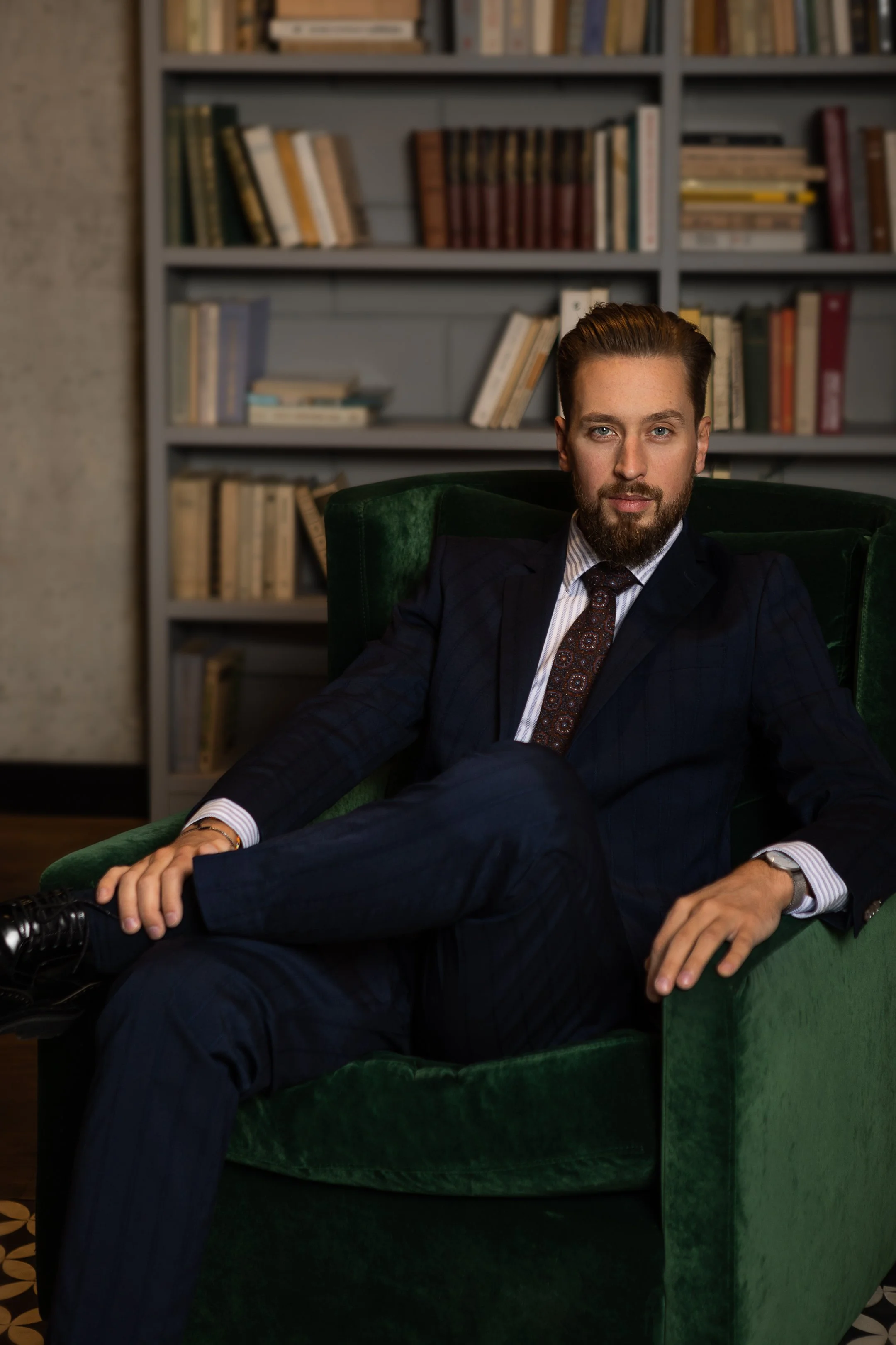 A man in a suit sitting on a green armchair in front of a bookshelf filled with books.
