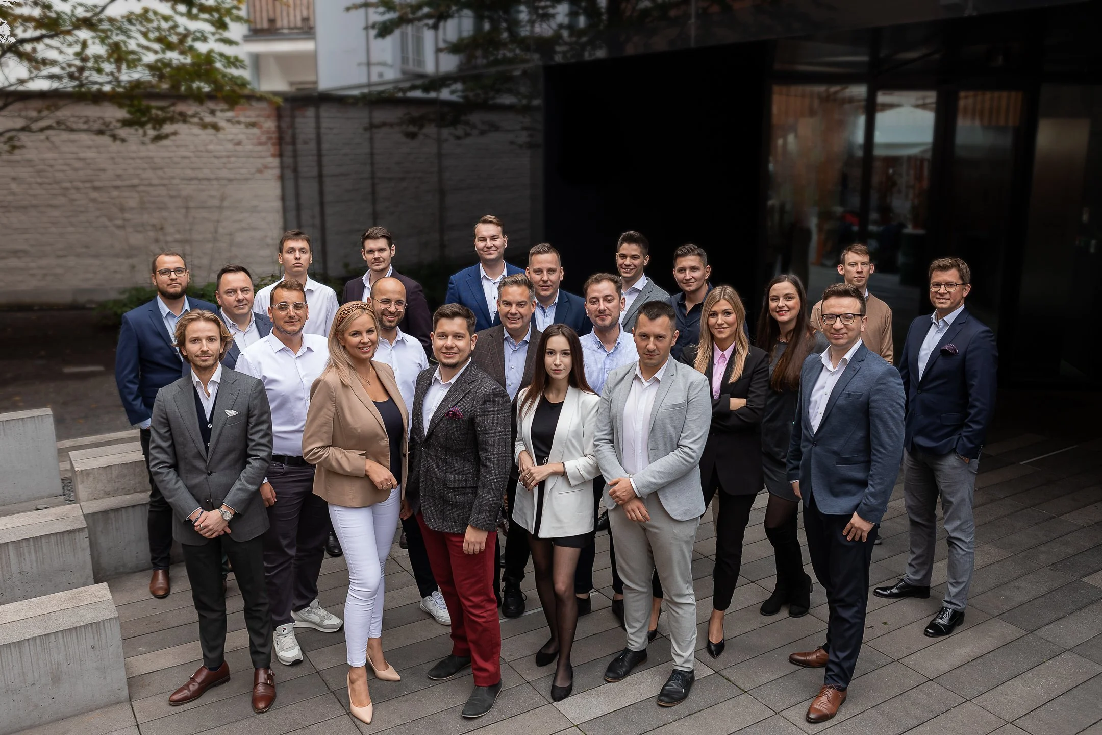A group of 21 professionally dressed men and women posing together outdoors, with a modern building and trees in the background.
