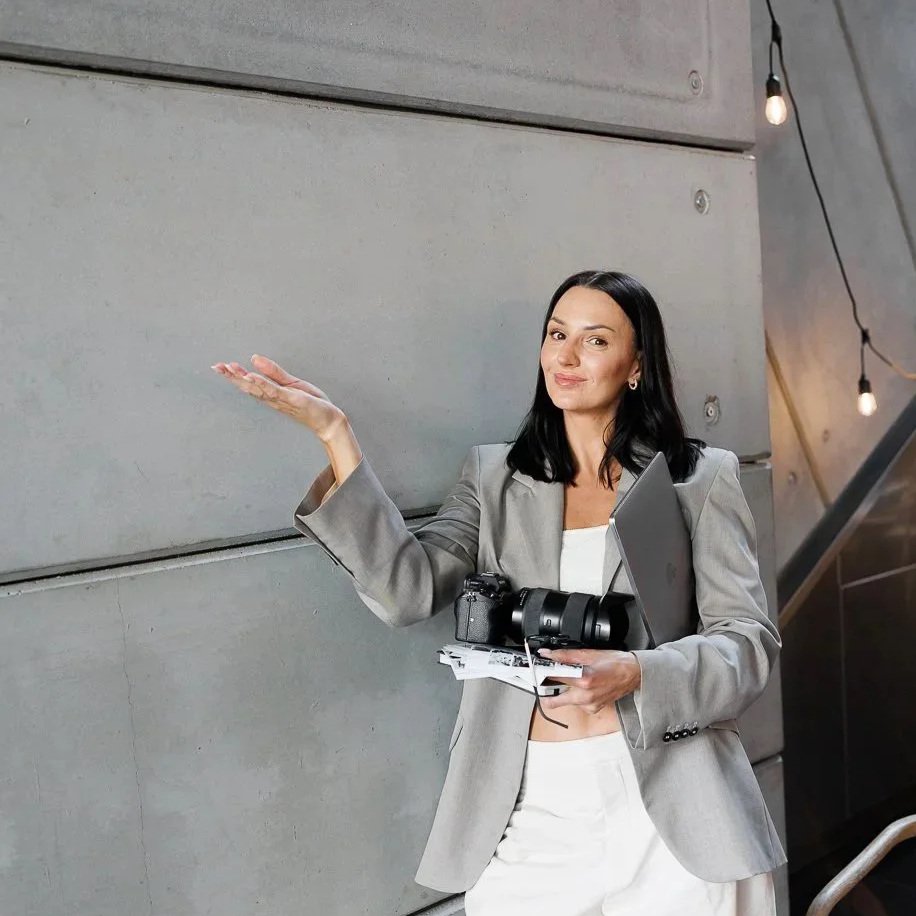 A woman in a gray blazer holding a camera, a laptop, and papers, gesturing with her left hand inside a modern building.