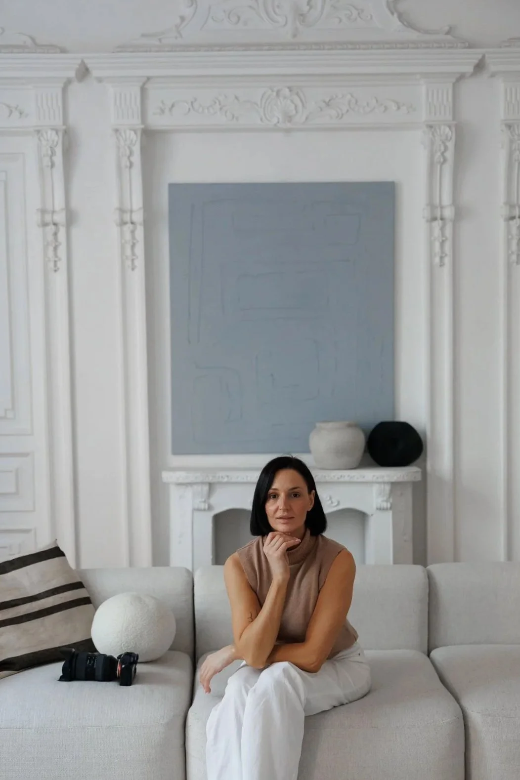 A woman with short black hair seated on a white sofa in a room with white ornate walls and a fireplace, with two decorative vases on the mantle behind her.