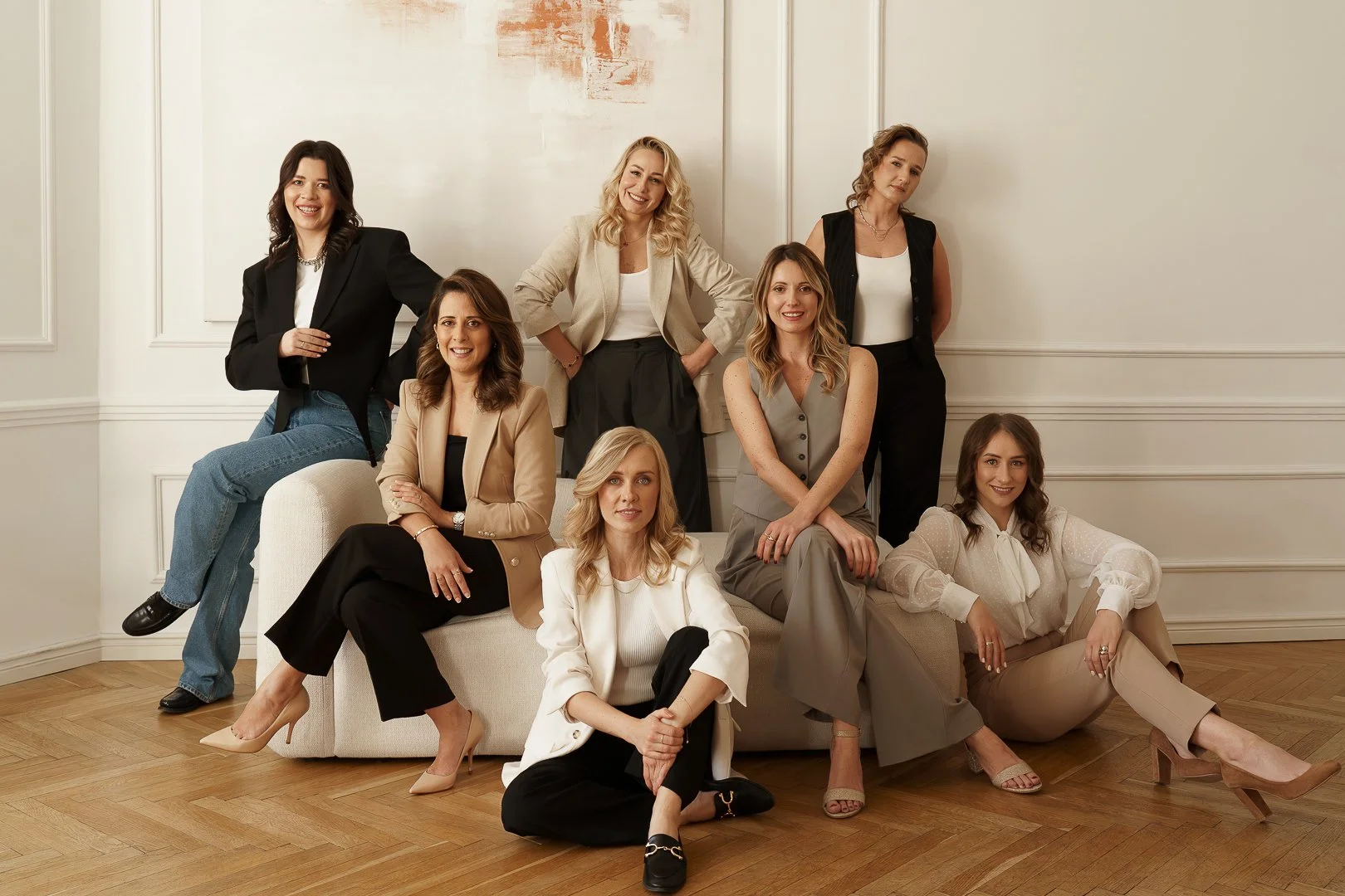 A group of nine women posing together in a well-lit room with white walls and wooden floors. They are dressed in business casual and formal attire, smiling and looking at the camera.