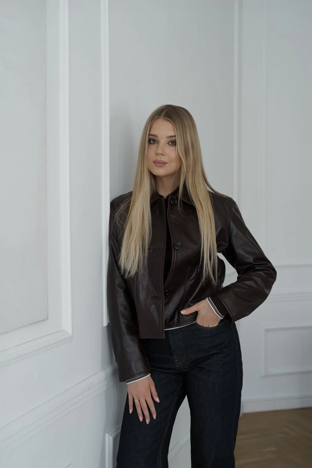 A young woman with long blonde hair wearing a black leather jacket and black jeans standing against a white wall with paneling.