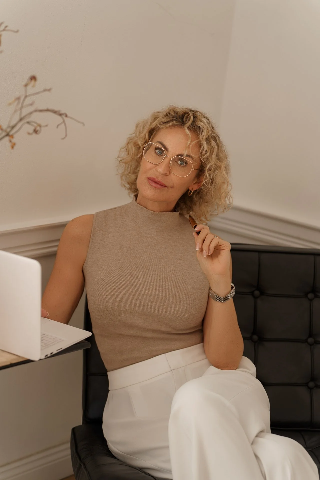 A woman with curly blonde hair and glasses sits on a black chair, holding a cigar and looking at the camera in a neutral expression. She is wearing a sleeveless beige top and white pants, with a watch on her left wrist. There is a laptop on a table b