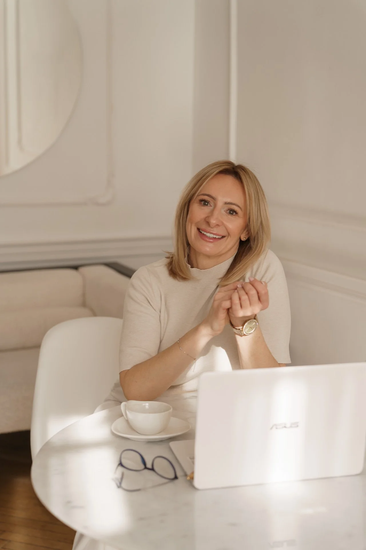 A woman with shoulder-length blonde hair smiling at a table with a laptop, a cup and saucer, and glasses, in a bright room with white walls.