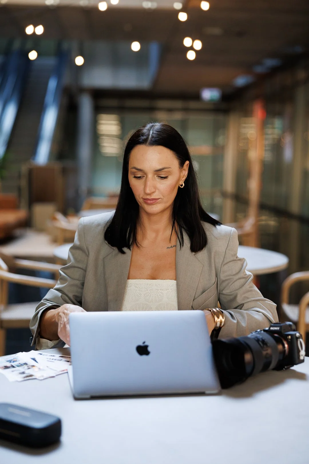 A woman with black hair sitting at a table with a silver MacBook, a camera, and some papers in a modern, well-lit indoor setting.