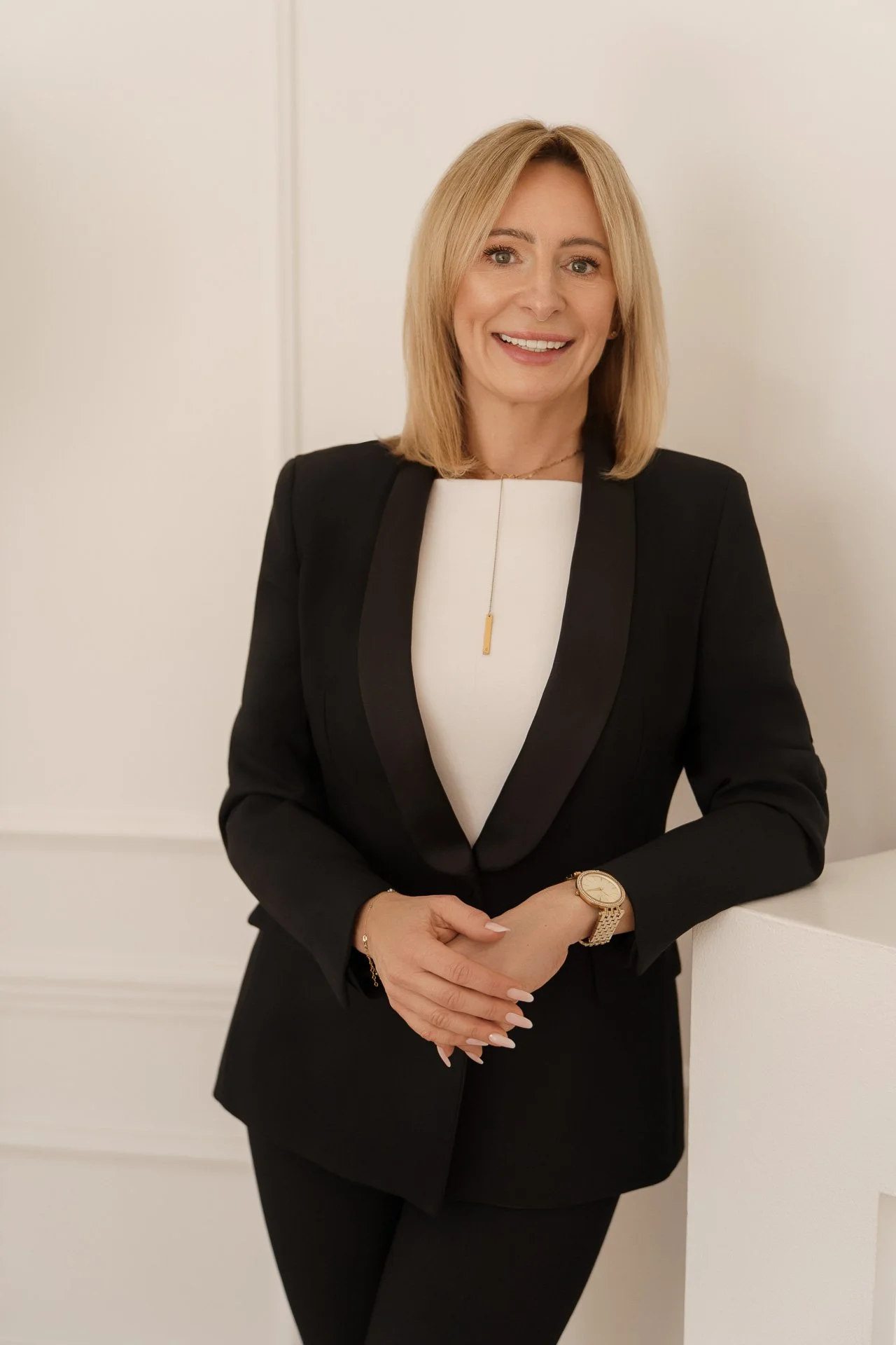 A professional woman with blonde hair, wearing a black blazer and white top, smiling and standing against a cream-colored background.