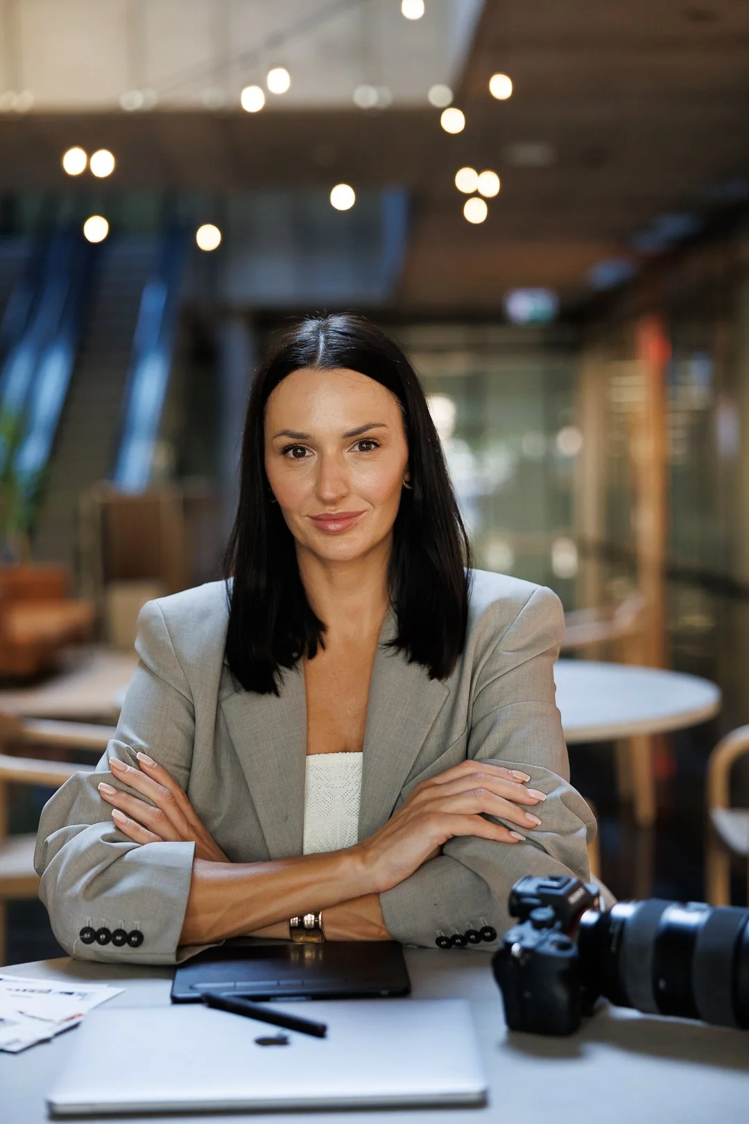 A woman with dark hair wearing a light gray blazer sitting at a table with a tablet, camera, and papers in a modern indoor setting with warm lighting.