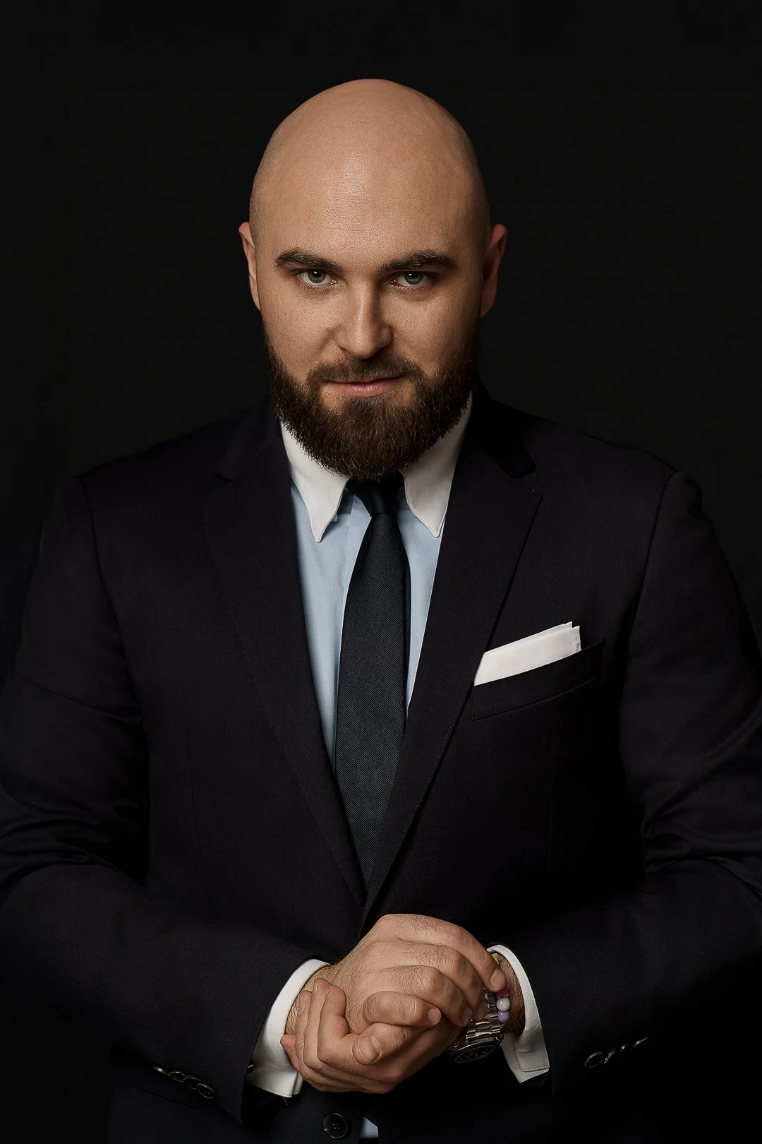 A bald man with a beard wearing a dark suit, white shirt, and dark tie, posing with hands clasped in front of him against a black background.