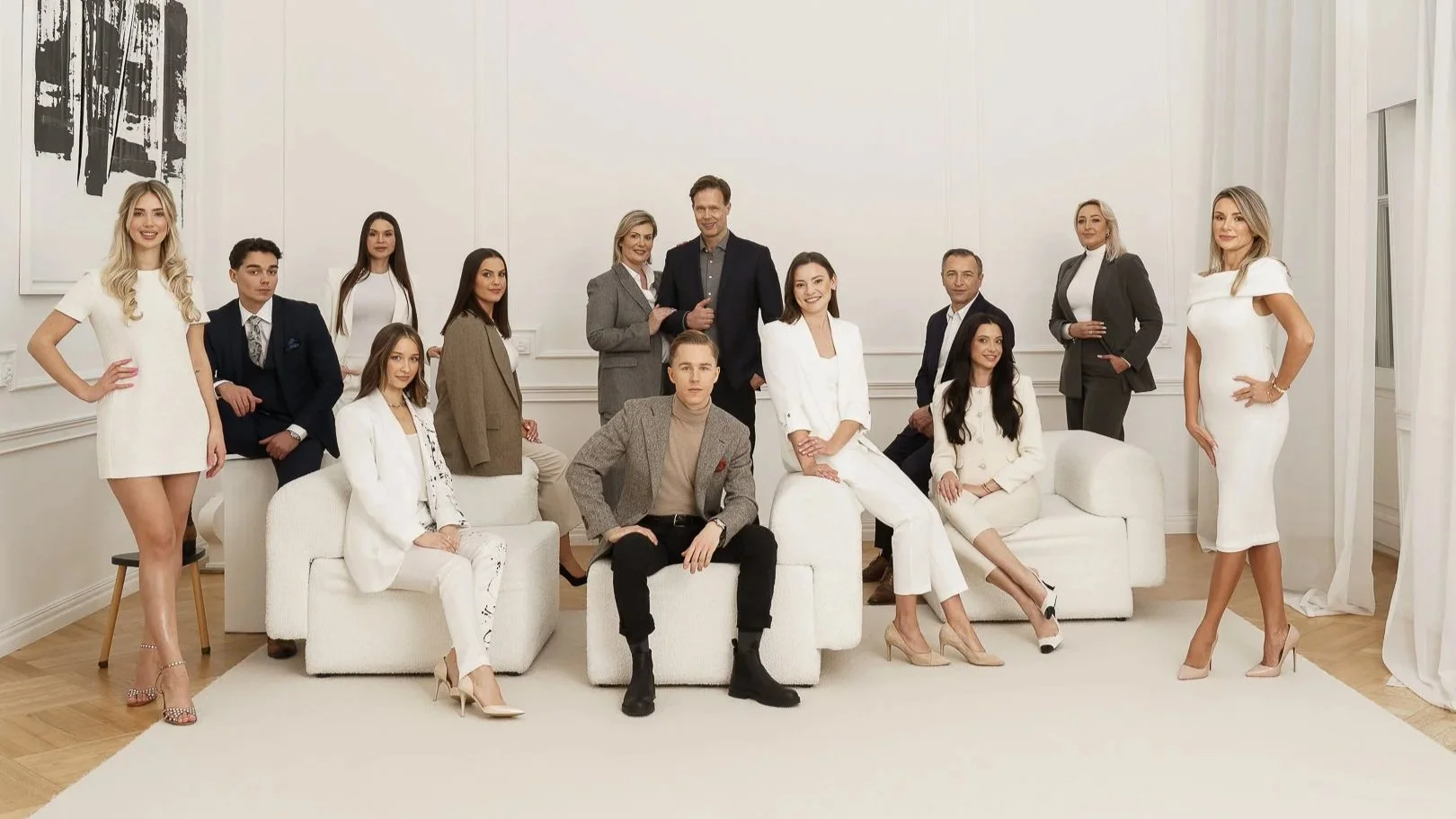 Group of fourteen professionally dressed people posing in a well-lit, elegant room with white walls and wooden floors.