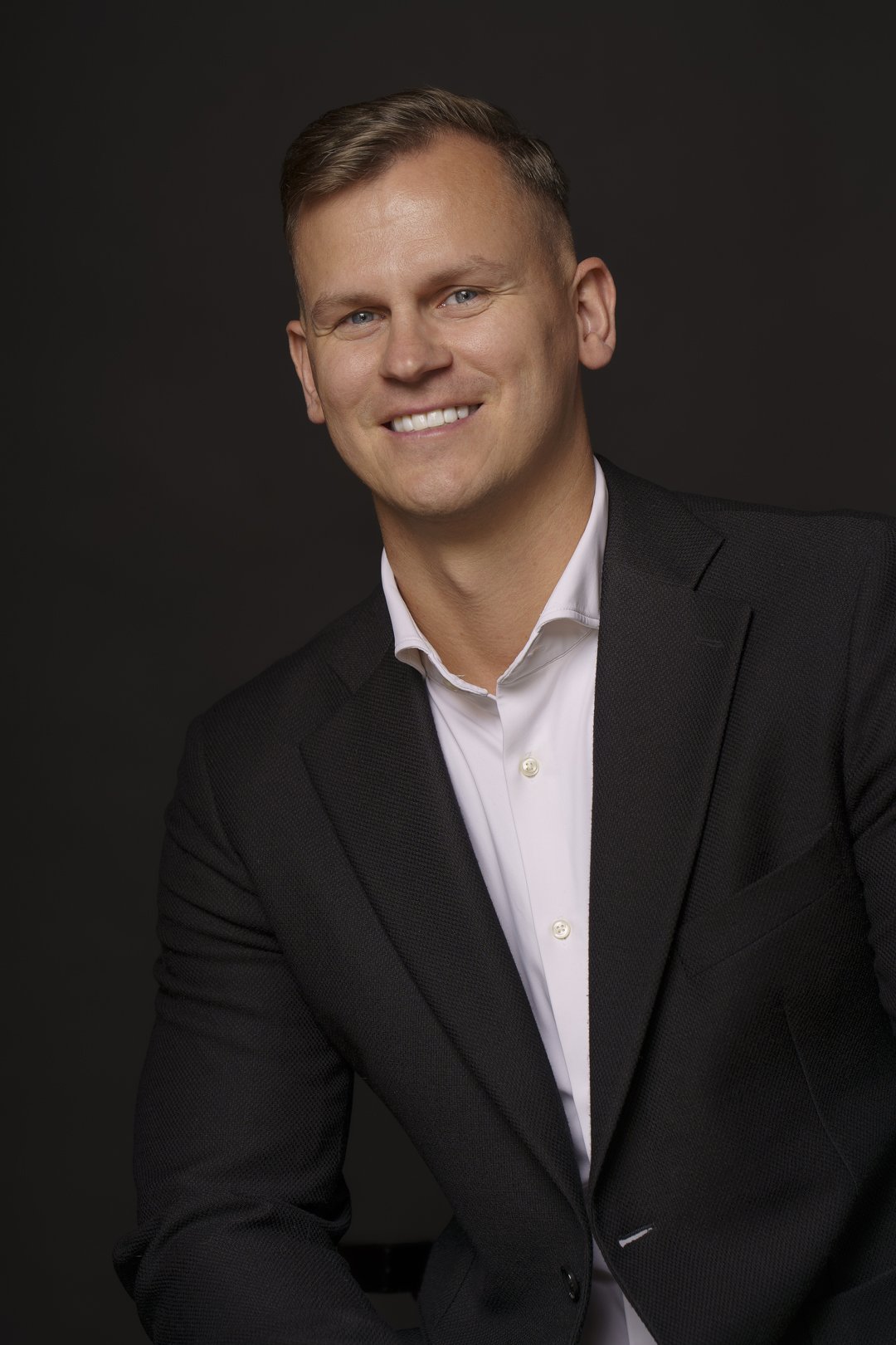 Professional headshot of a smiling man in a black suit and white shirt against a dark background.