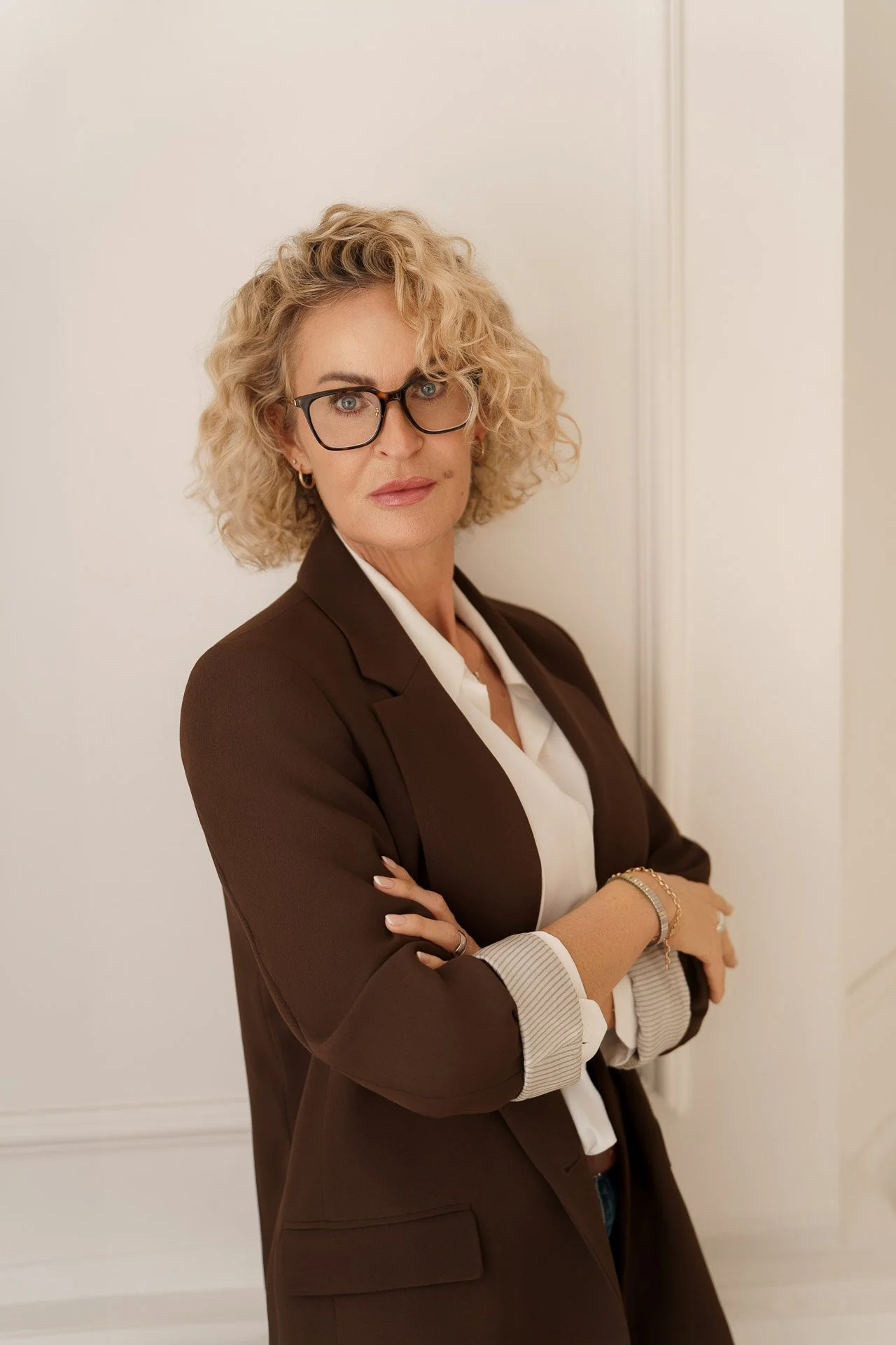 A woman with curly blonde hair and glasses stands with her arms crossed against a white wall, wearing a brown blazer and a white blouse.