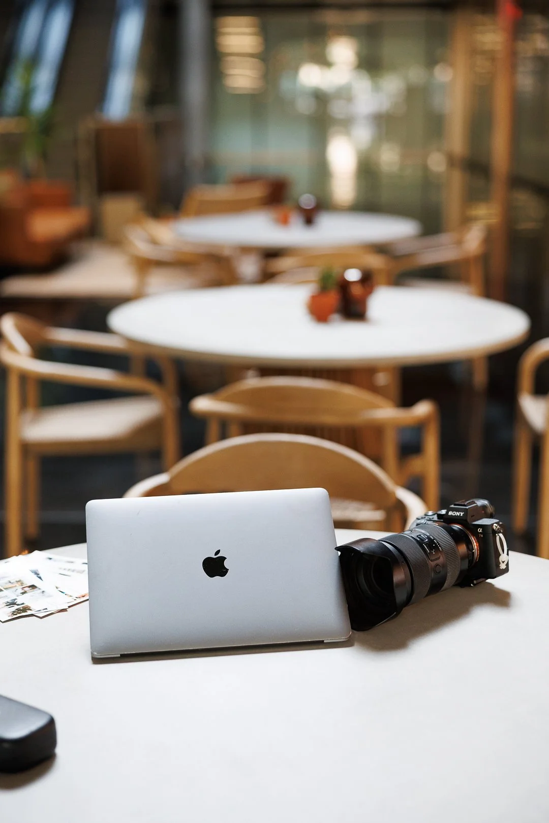 A closed silver MacBook, a black camera with a large lens, and some small photographs on a white table in a bright, modern cafe with wooden chairs and round tables.